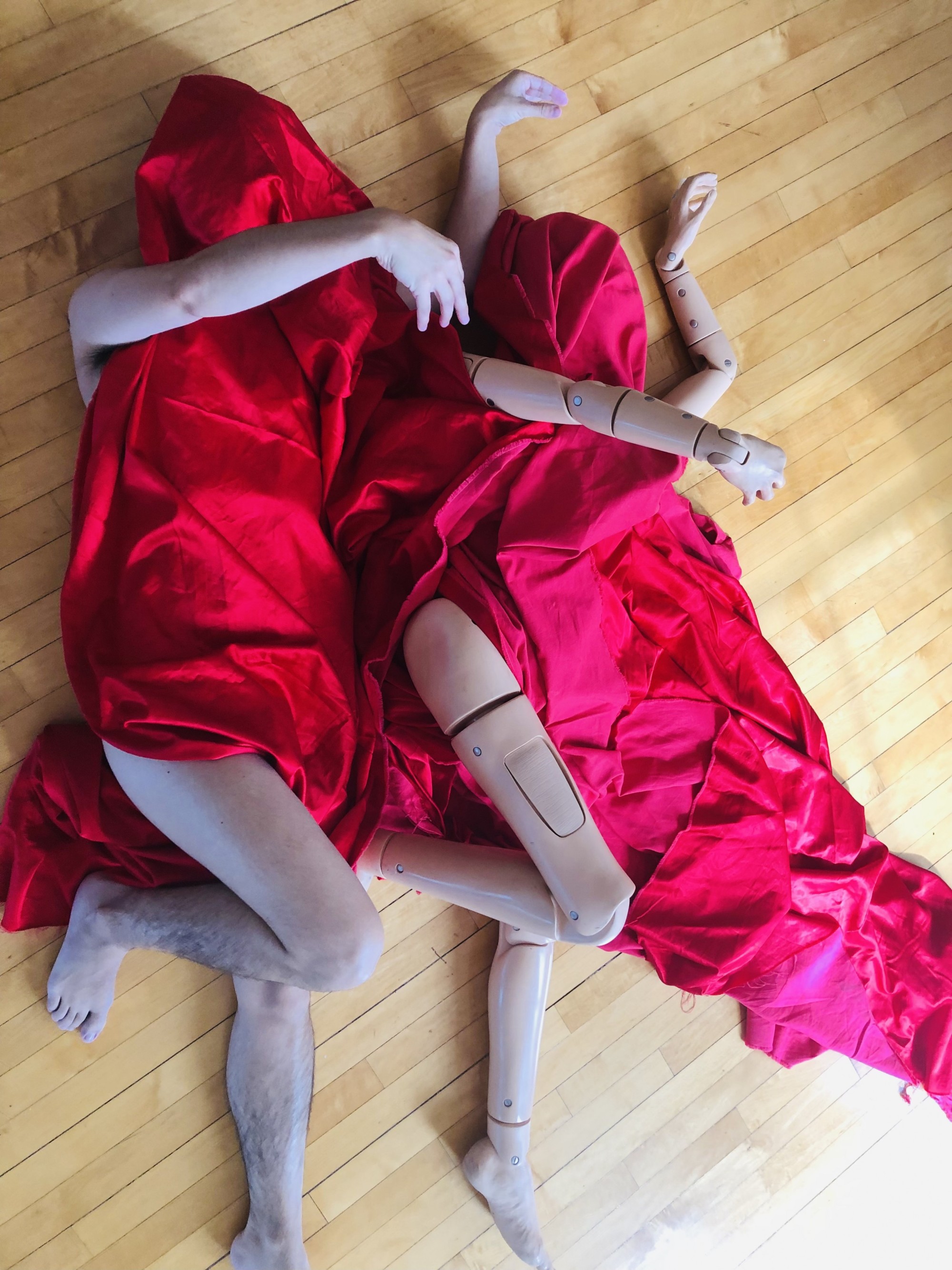 Two figures are lying on a wood floor, draped in a red blanket that covers everything but their arms and legs, which are positioned identically to each other. One figure is devynn emory, are the other is Cindy, a medical mannequin