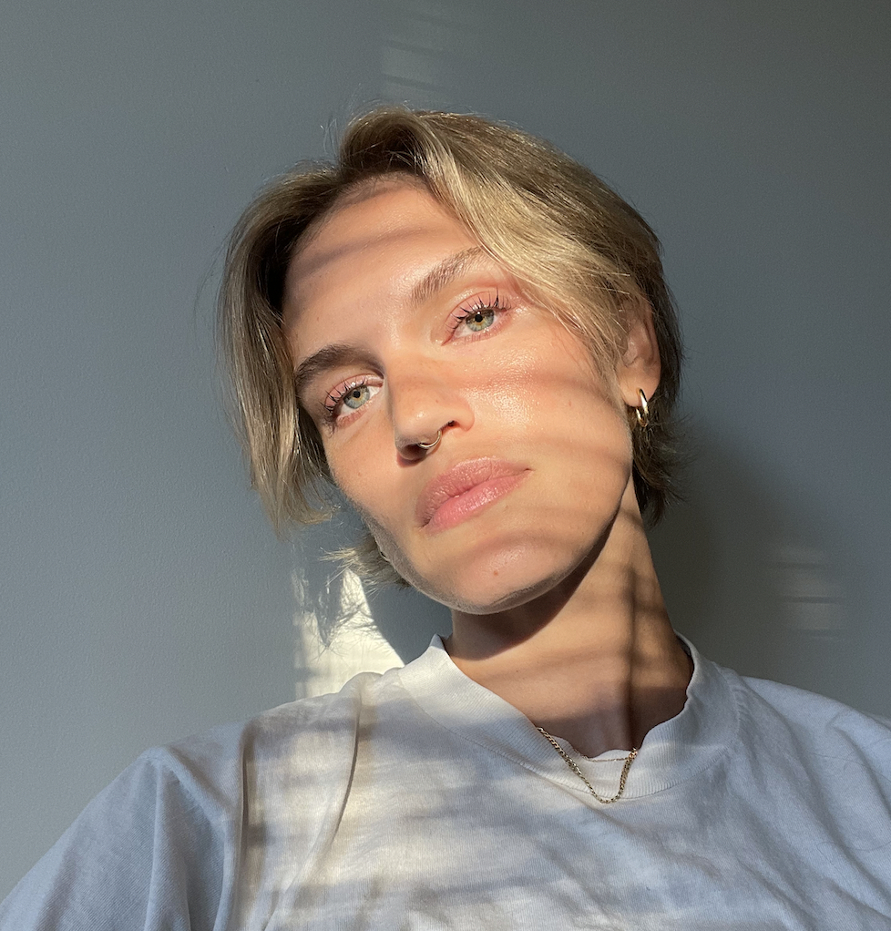 A person with short blonde hair is seen from the shoulders up against a white wall.