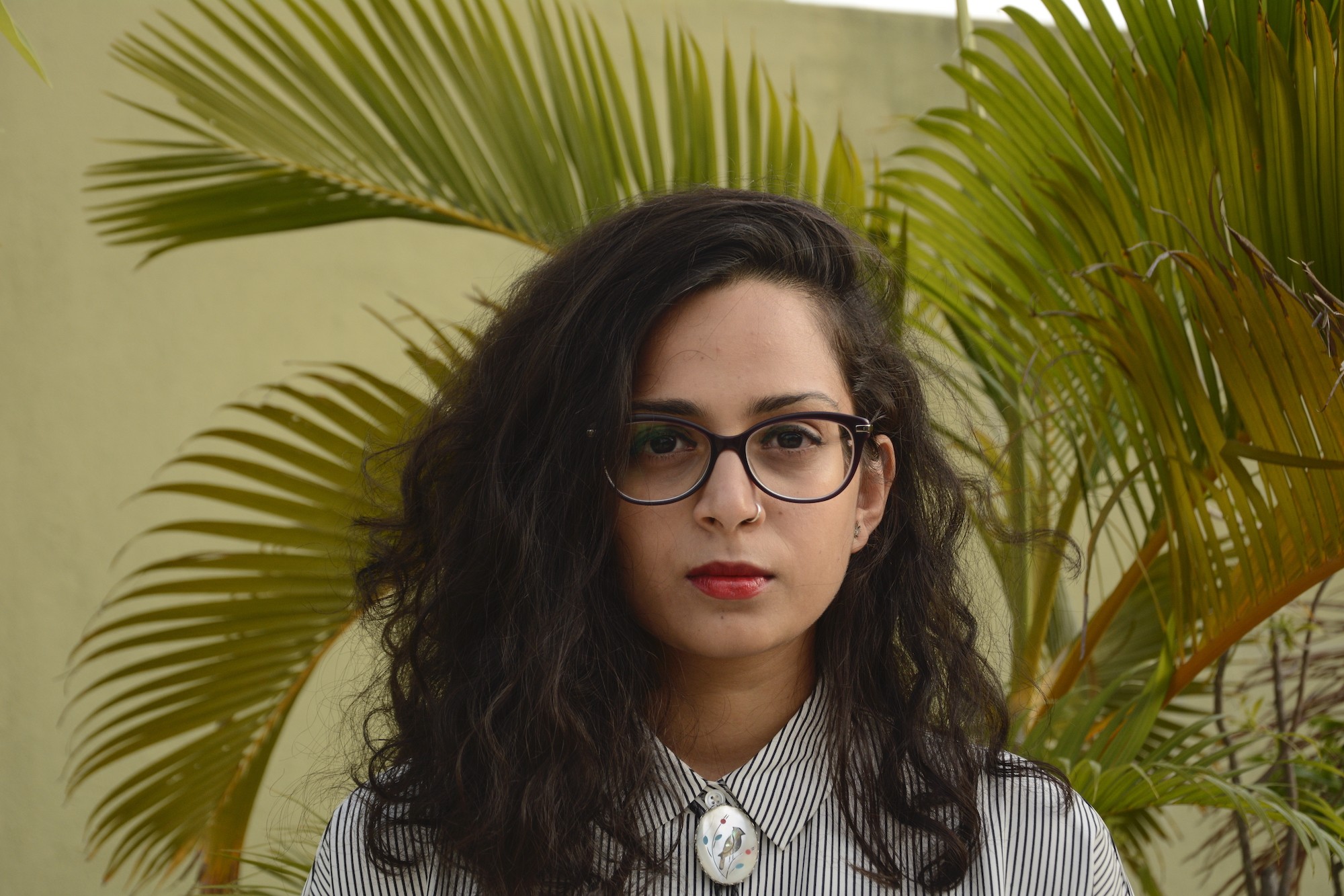 Aditi Machado stands in front of an areca palm, wearing red lipstick and black frame glasses.