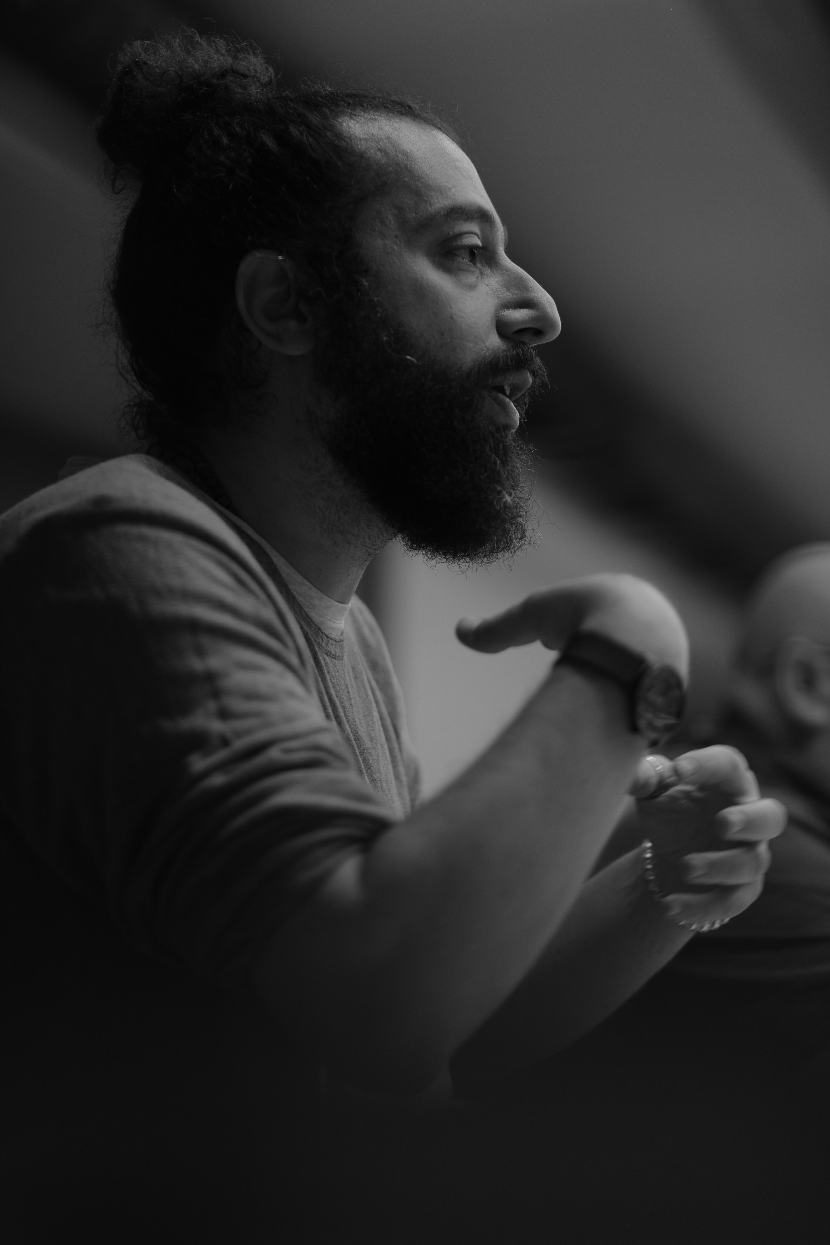 Ahmad Alhmallah is shown in a black-and-white portrait-oriented photograph, cropped from the torso up. He is shown in profile, his long hair up in a bun. He is wearing a t-shirt and gesturing with his arms as the photograph captured him mid-speech.