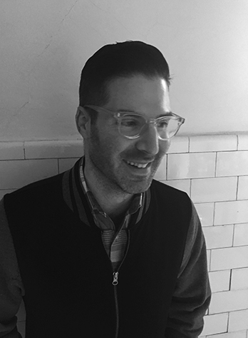 Black and white photo of Alan in a black jacket, button up shirt, and glasses smiling against a tile wall.