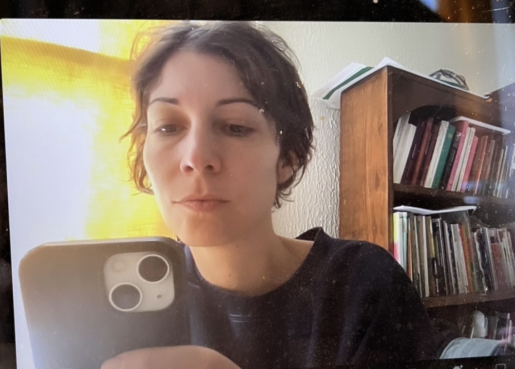 Alexis sits at her desk next to a yellow curtain, holding the phone up to take a selfie before a zoom meeting starts. She's wearing a navy blue sweatshirt, and there's a bookshelf behind her filled with books.