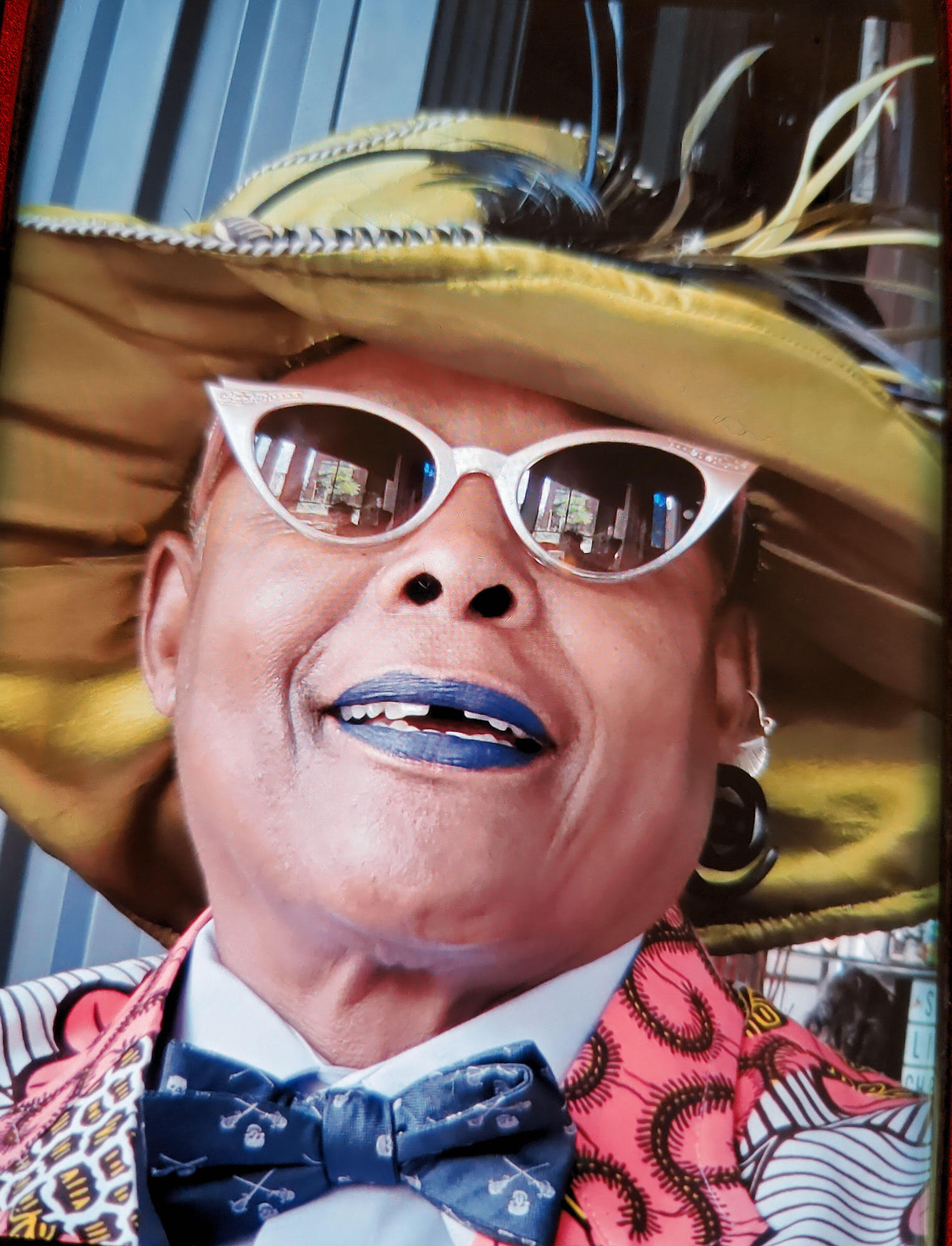 Alexis appears in a portrait-oriented color photograph, cropped tightly around the head from the neck up so that almost no background is visible. She is wearing a brightly colored, multi-patterned vest or jacket, a white button down, and a dark blue bowtie with small skulls and crossed swords on it. She is wearing a fantastic yellow or chartreuse hat with multicolored feathers, silver metal cat eye sunglasses, and a blue matte lipstick that almost matches the blue of her bowtie. She is looking up and to the left and smiling.