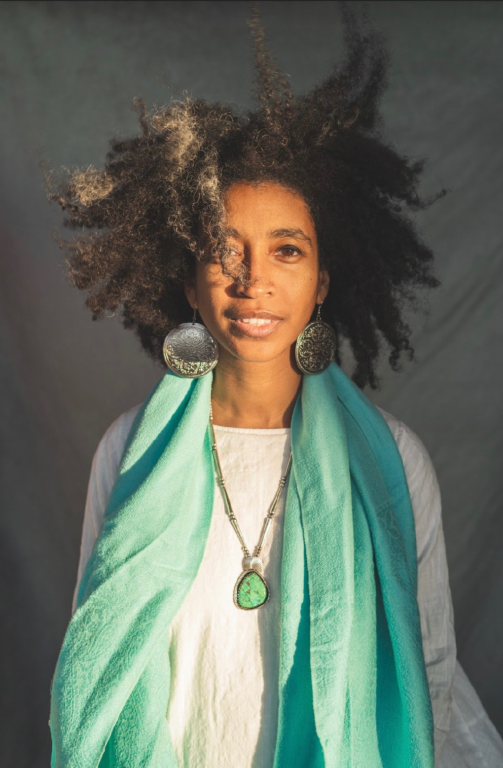 Alexis Pauline Gumbs stands in the sun wearing a white shirt and blue-green scarf, her/their hair lifted by the wind