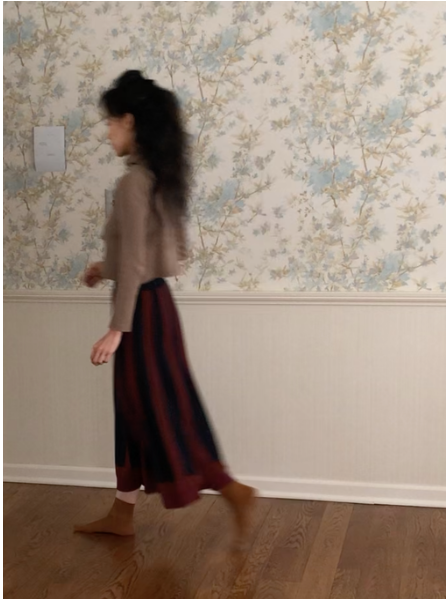 Ali walks across a wooden floor, in front of blue, green, and white floral wallpaper. She wears a brown shirt, red and black skirt, brown socks, and has long, dark brown hair. Image is blurred.