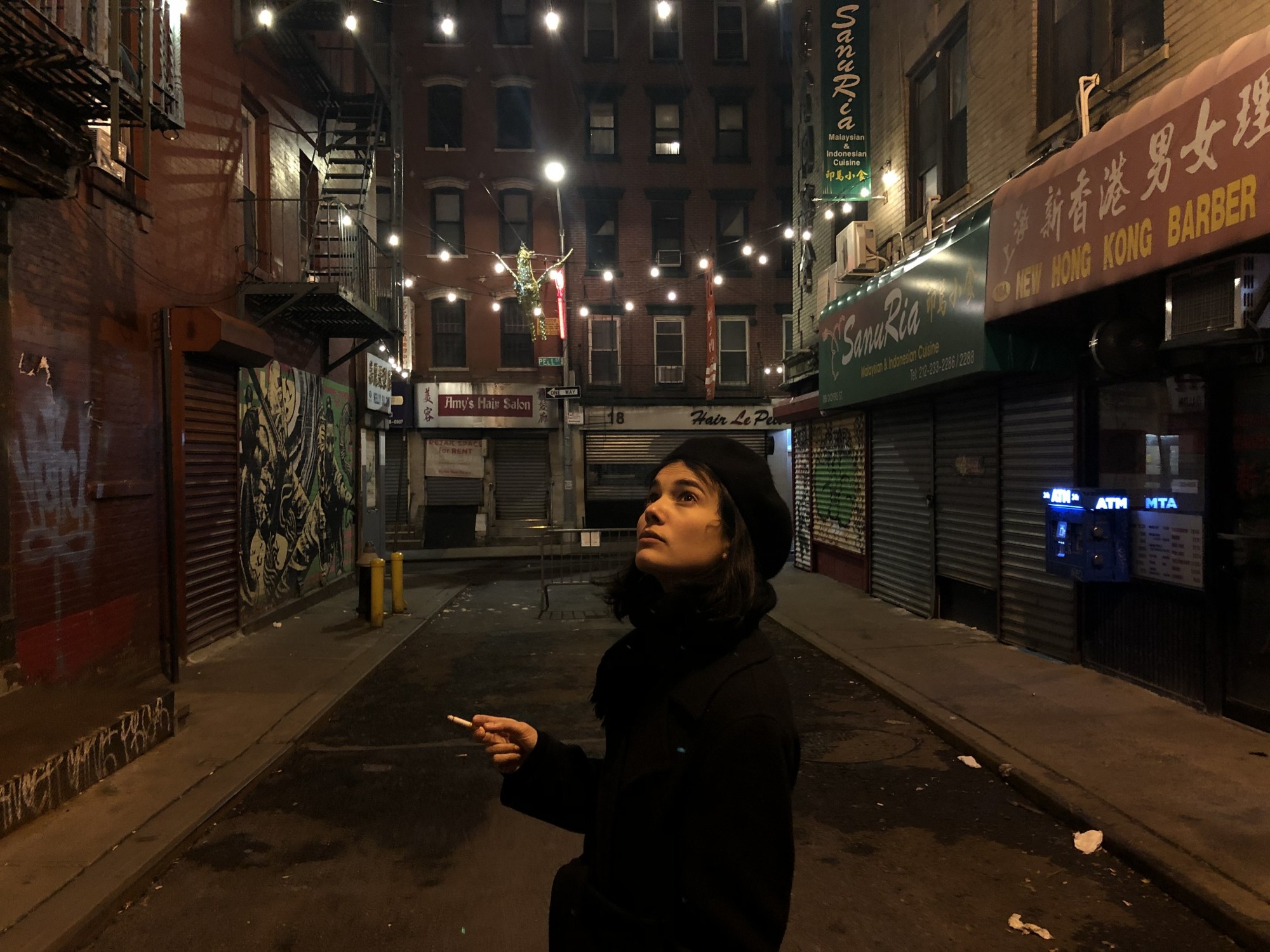 Alisha Mascarenhas stands in the middle of a deserted nighttime street in Manhattan, smoking a cigarette and looking up at the lights that hang between the buildings