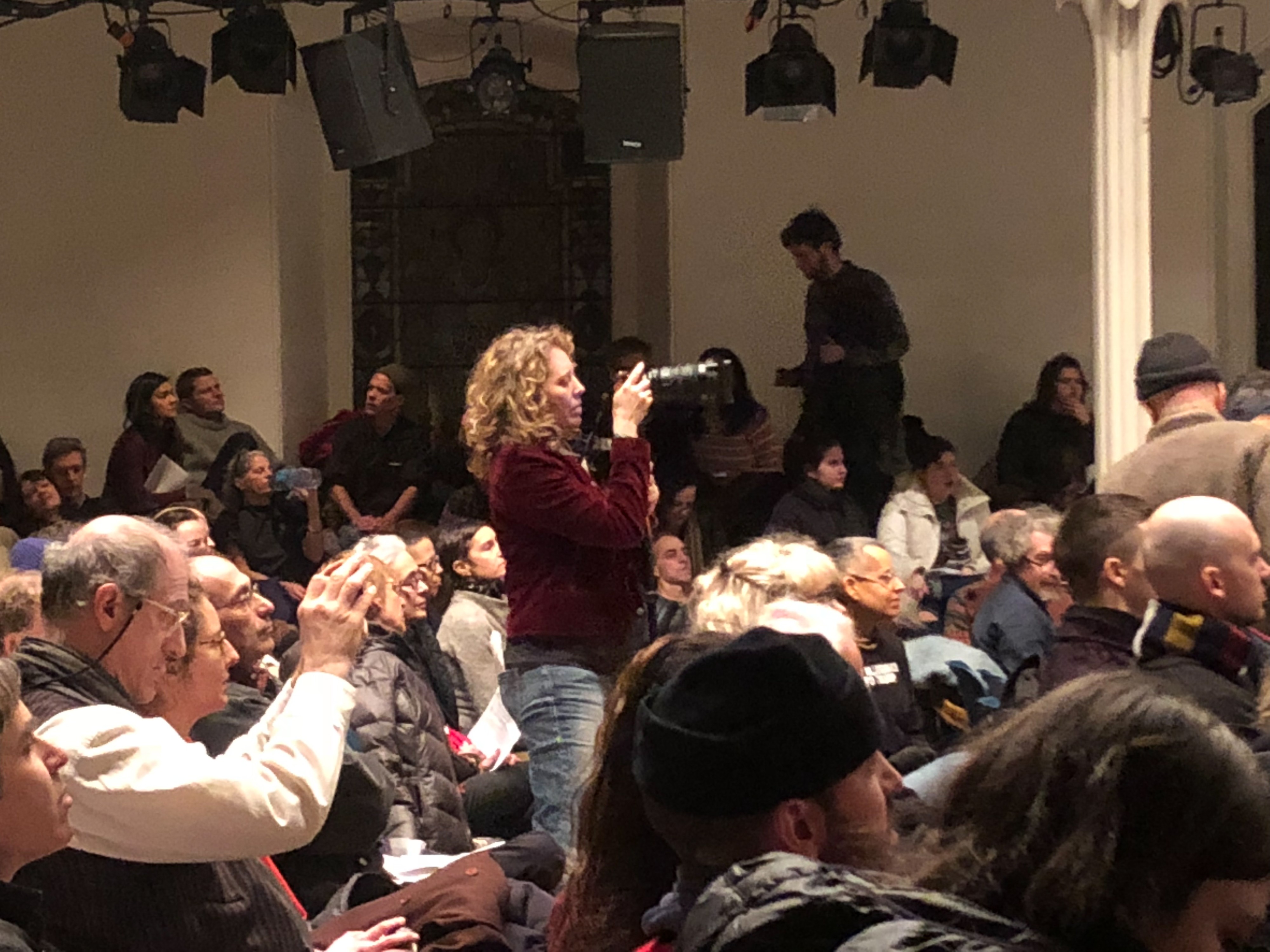A blonde woman with curly hair stands amid a large crowd, pointing her camera toward the stage.