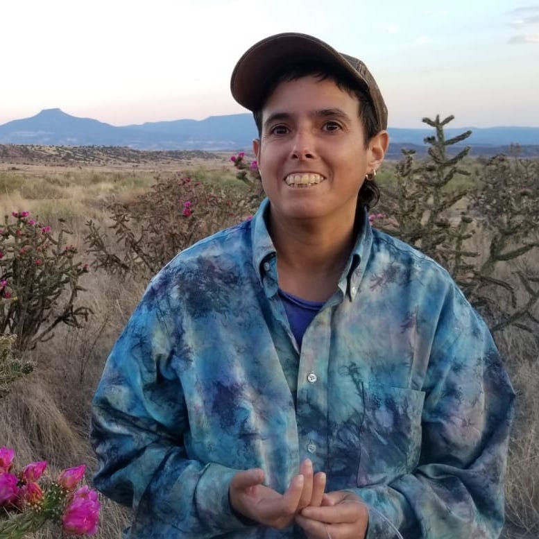 Amelia is centered in a square photograph, cropped from the waist up. Wearing a blue tie-dyed button down and brown-green hat, she is smiling and facing the camera. Behind is a desert landscape with brush and desert foliage. Mountains are visible in the background.