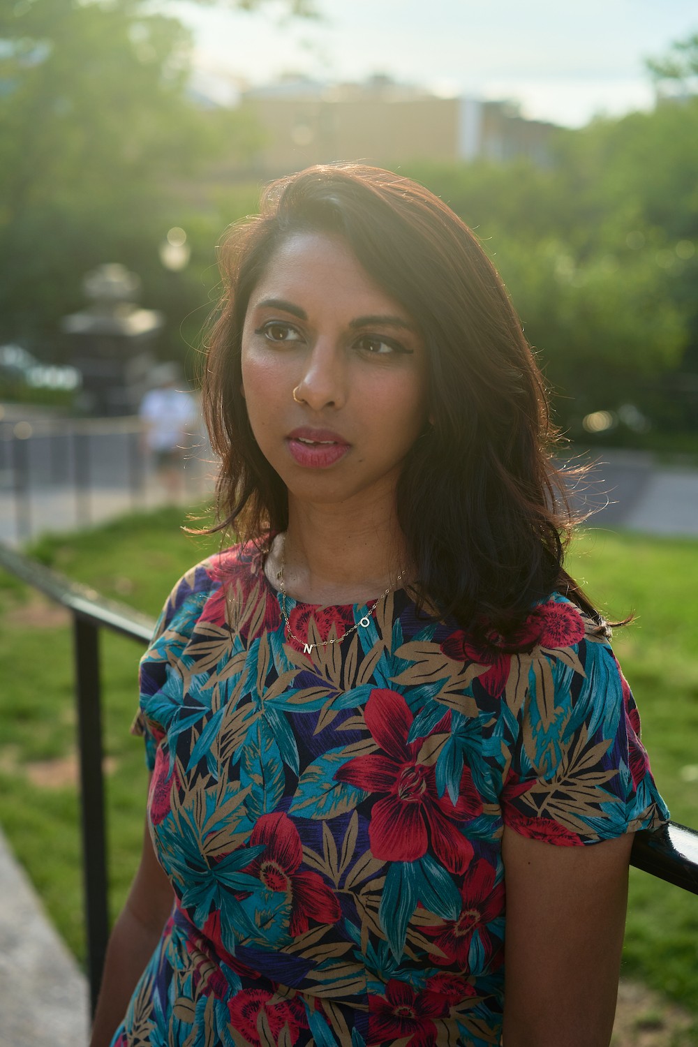 Amirtha Kidambi is featured centered in a portrait-formatted photograph, looking off to the left side of the photograph. She is wearing a floral dress and has shoulder-length hair. Behind her is a grassy landscape resembling a park. The photograph is suffuse with bright sun.