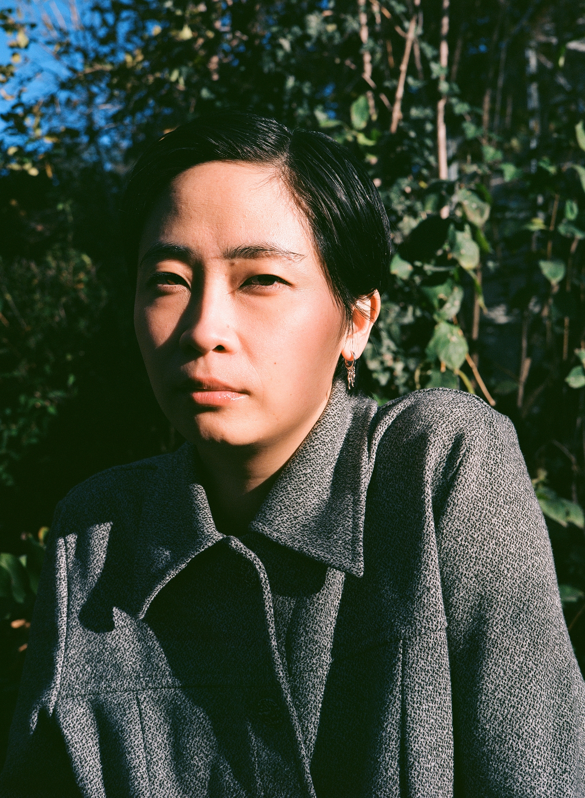 Amy, an East Asian person with short black hair, is wearing a grey button-up top. She is sitting outside in front of a leafy green tree, with the left side of her face in shadow and the other side in the bright sun.