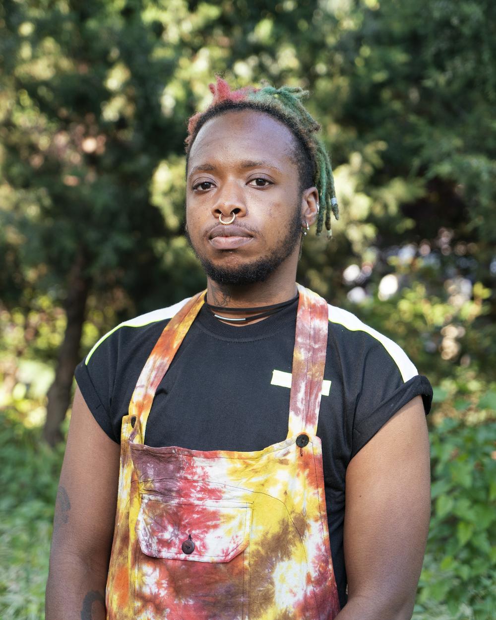 Anais stands in front a some greenery wearing overalls over a black t-shirt. He is looking at the camera