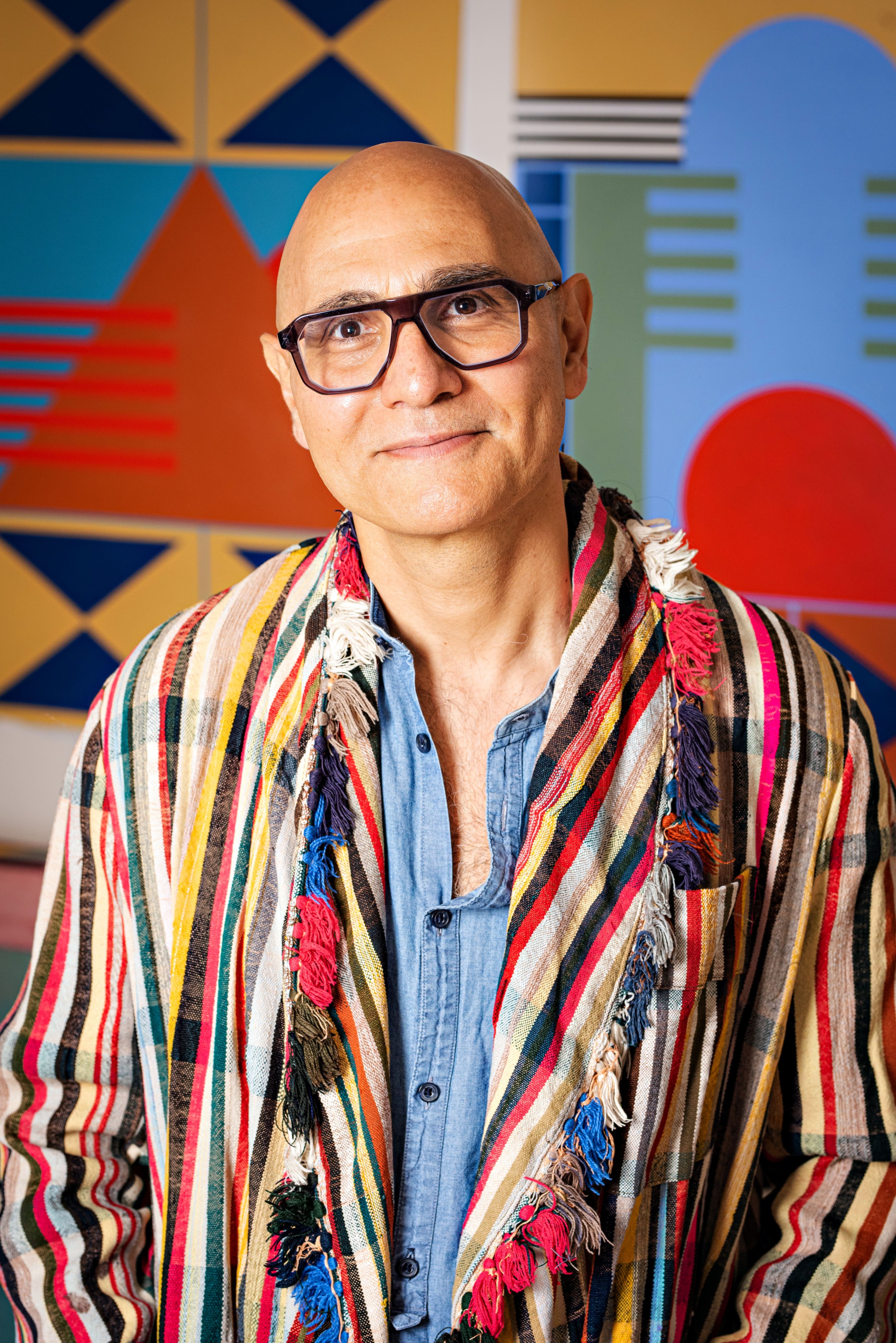 A bald person wearing black-framed glasses and a colorful striped jacket over a denim shirt stands before a patterned, vibrant background, smiling.