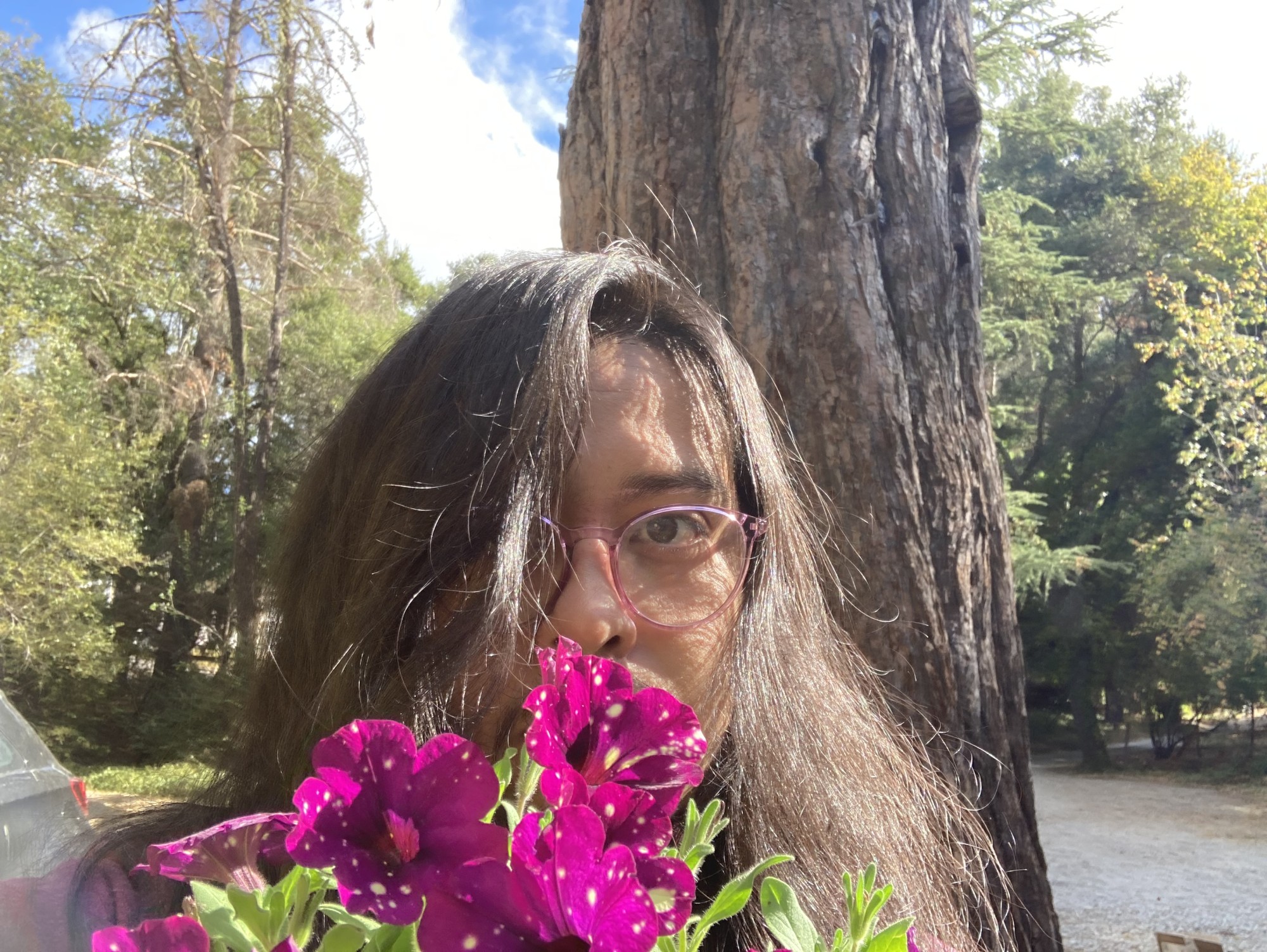 Angel stands outside by a large tree, their long hair framing their face, which is mostly obscured by the flowers they hold