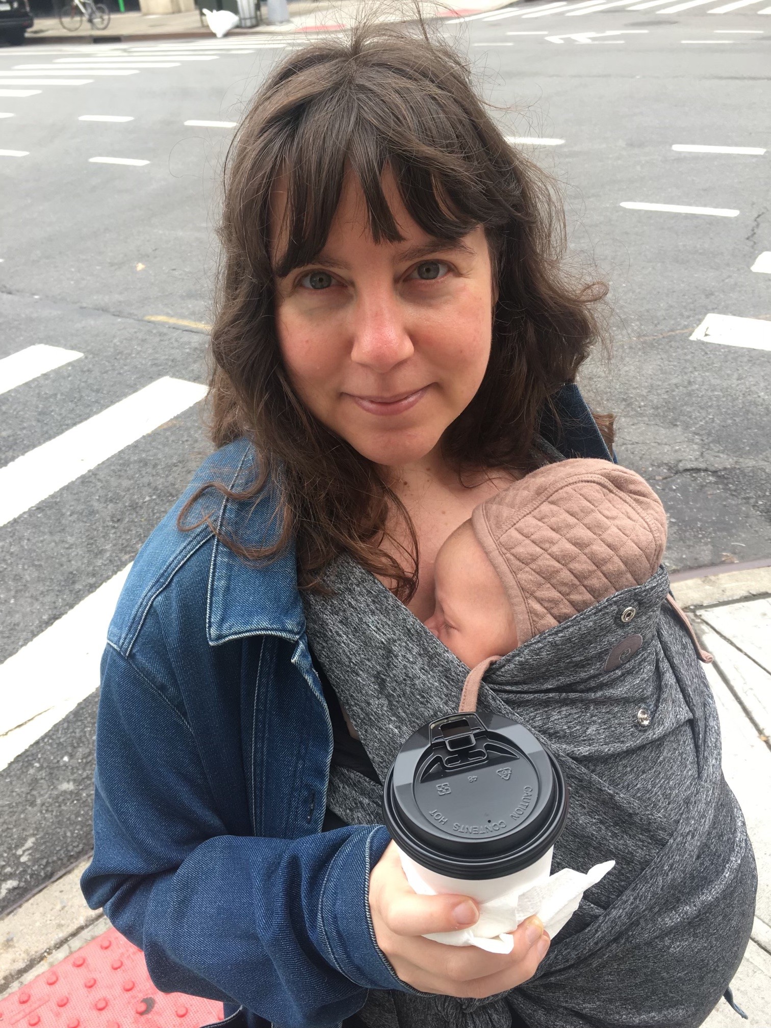 Anna is shown in a portrait-oriented photograph cropped from the waist up. The picture is taken at a slight angle looking down. She is holding a takeaway coffee cup in one hand. There is a baby strapped to her chest in a grey swaddle. Anna is wearing a denim jacket and her dark hair is loose and extends to her shoulders. The baby is wearing a quilted cap. The photo is taken at a street corner.