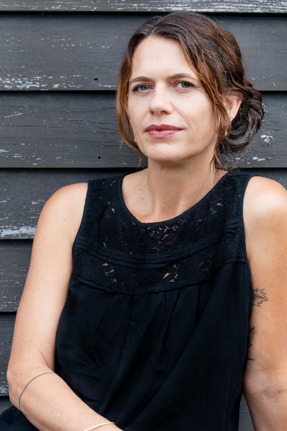 A white woman with olive skin wearing a black sleeveless shirt sits in front of a sunlit exterior house wall with black, chipping paint. She is looking at the camera and looks relaxed with a slight smile. Her dyed reddish-brown hair is loosely gathered and streaked with gray, and a fragment of a tattoo of a hawthorn plant is visible on her arm.