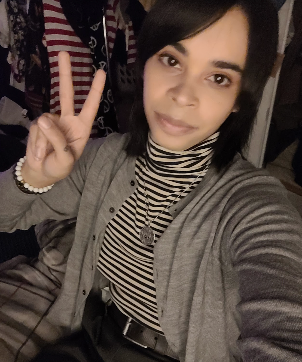 Slightly from above, a slightly smiling brown-skinned woman with a shoulder-length mullet in a gray cardigan, an off-white & navy-striped turtleneck, & a dark gray skirt with a black belt poses with a v-fingered peace sign for the camera. She also wears a black & white beaded bracelet. On her silver necklace is the sigil for the Buddhist deity Vajrayogini.