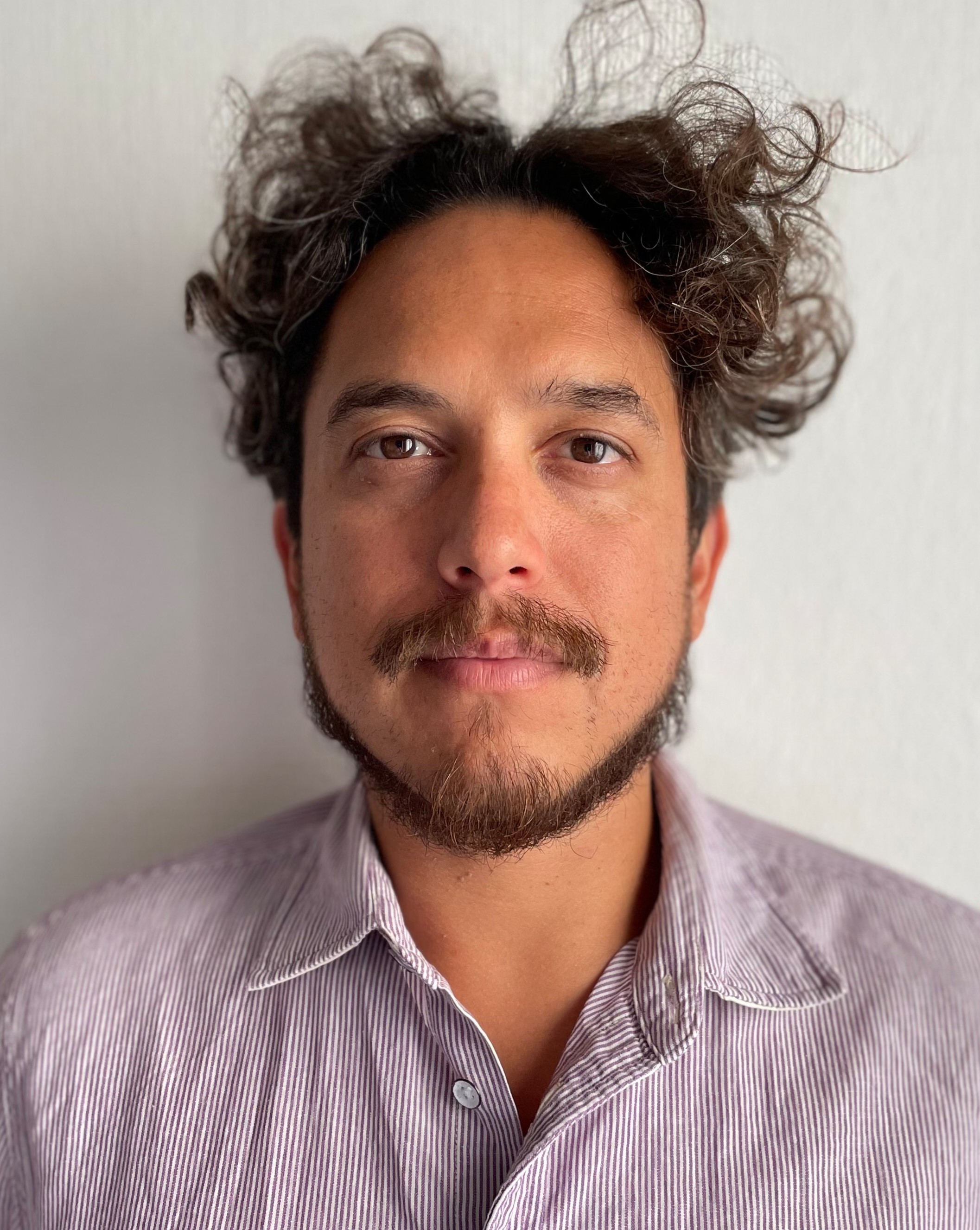 A person with short curly hair, a mustache, and a light beard stares at the camera, wearing a striped long-sleeve shirt against a white background.