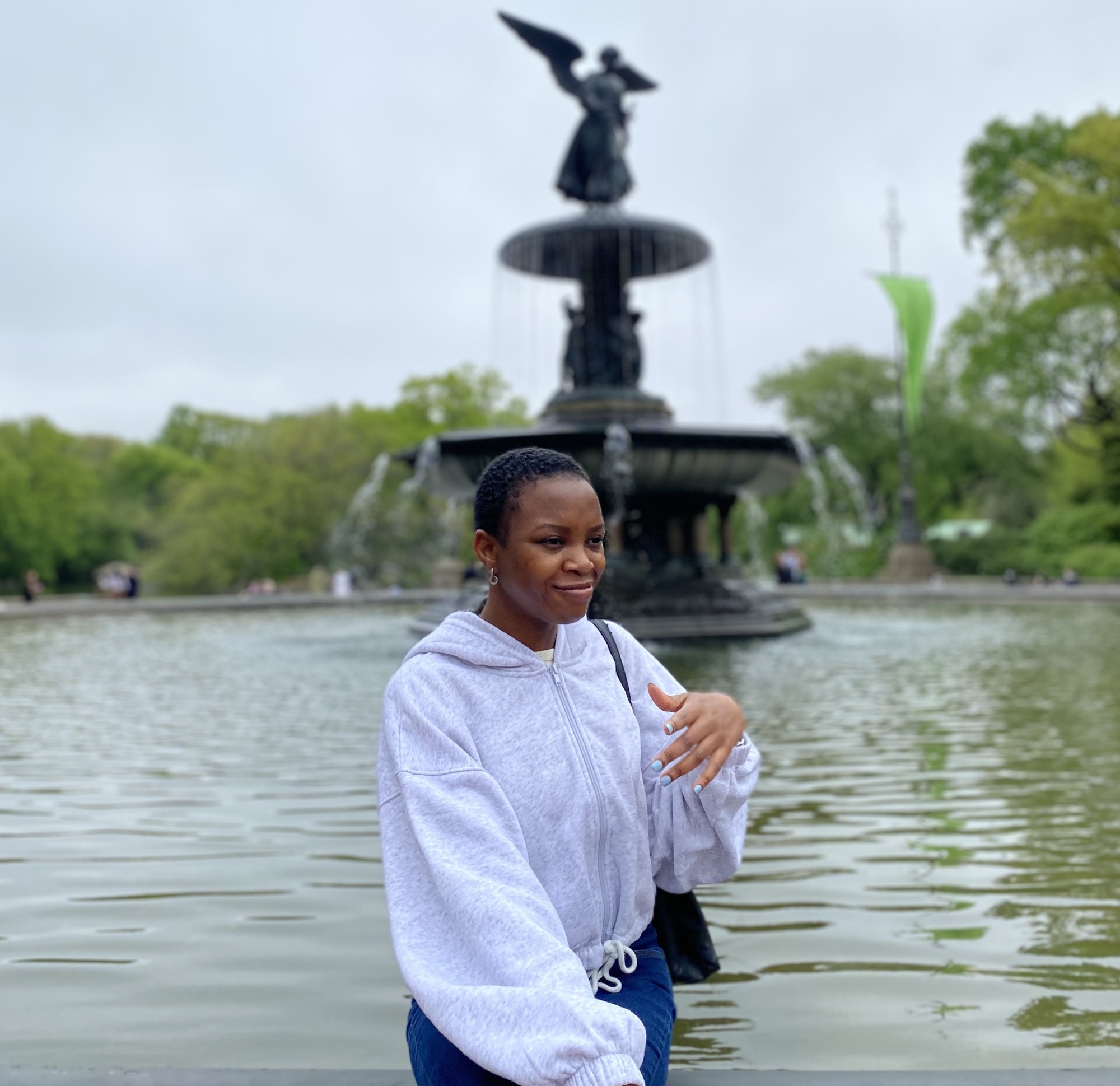 In this color portrait, Bibiana Ossai stands in front of a large fountain featuring a winged angel out of focus in the background. Ossai wears a grey zip-up hoodie and looks to the side