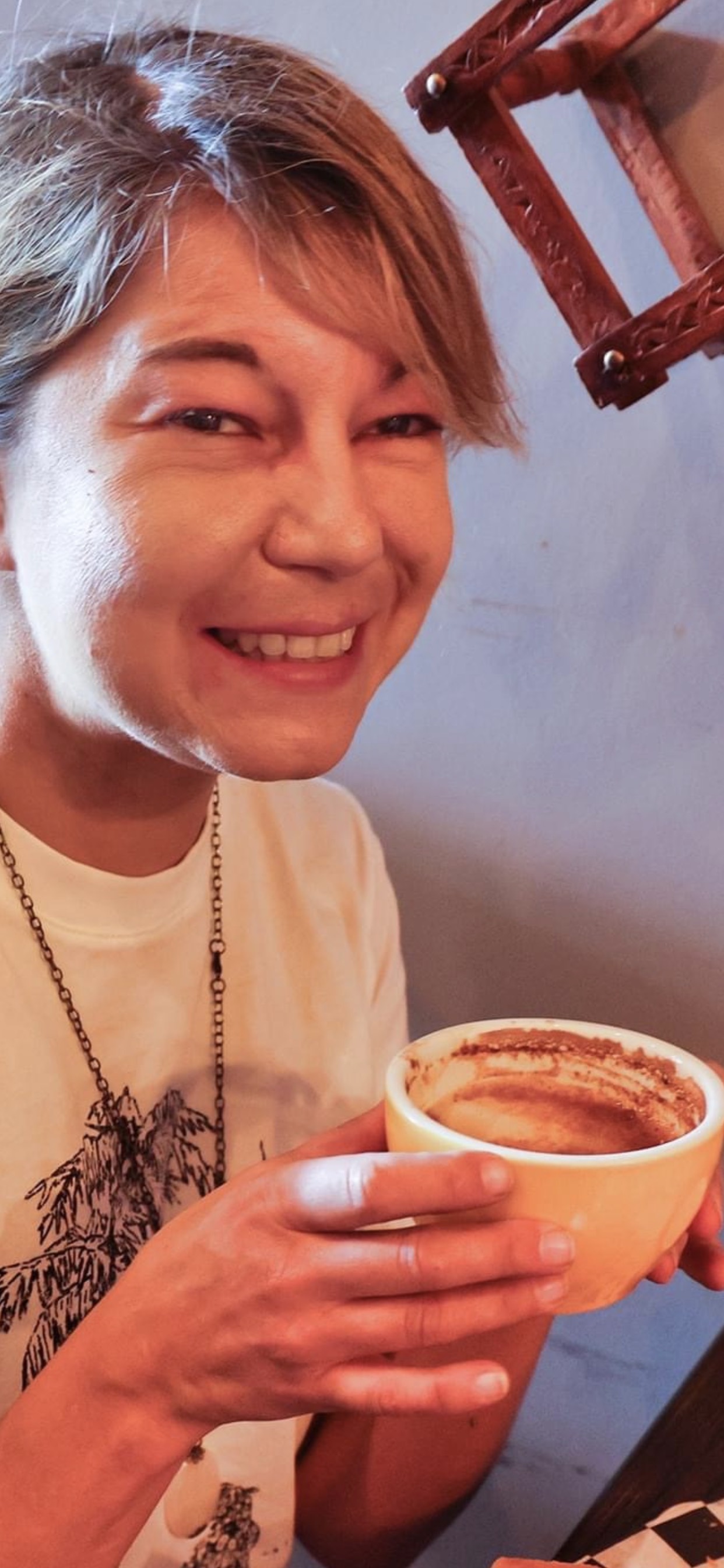 Bojana, a smiling white woman in her 40's, holding a fresh, hot, cup of coffee - as if to say, come join me for a chat. Her blond hair is pulled back into a pony tail. Her bangs are swept to the left side, and she is wearing a soft, cream colored, button down shirt.