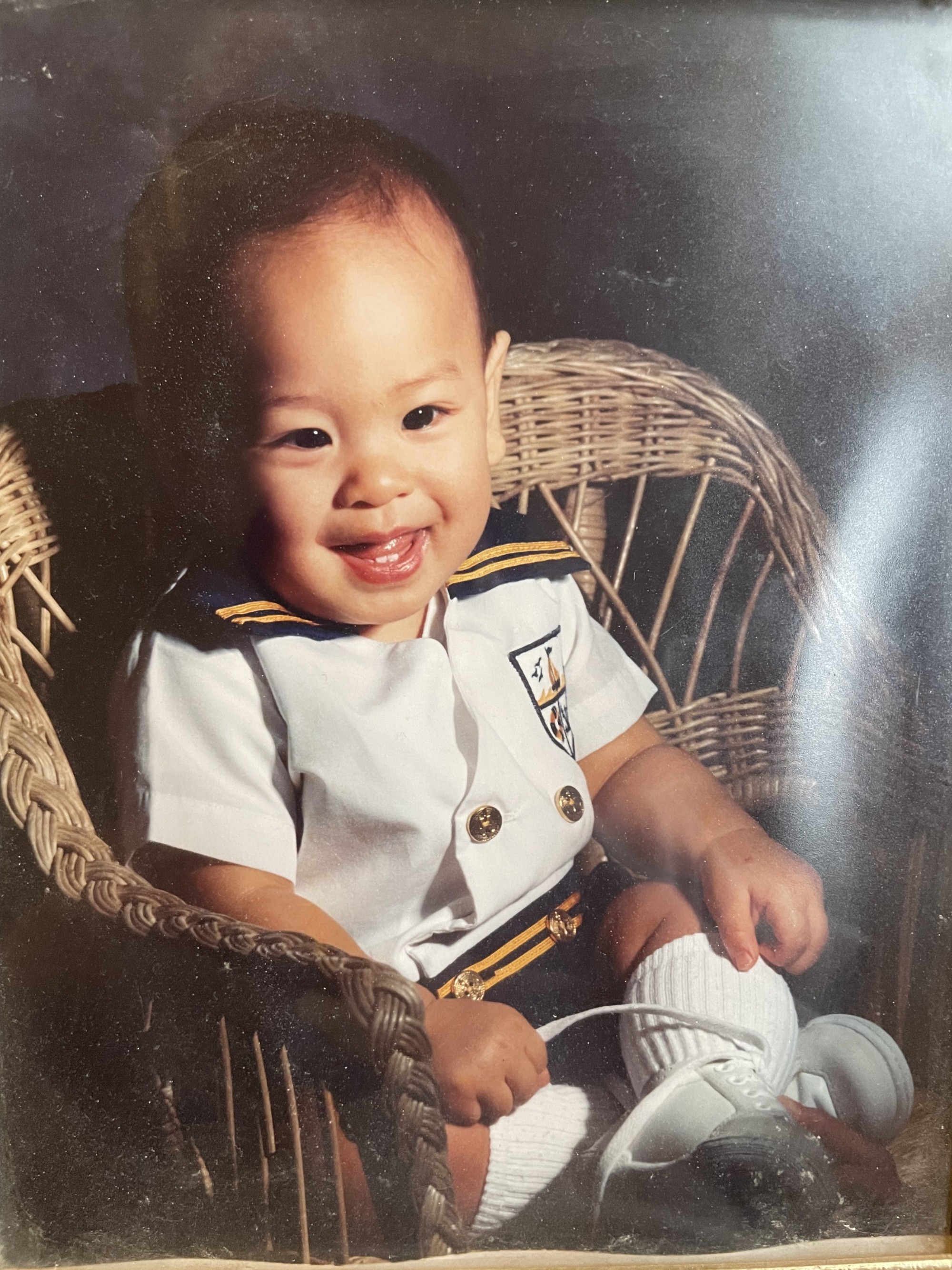 Cean, a Filipino baby with a sailor outfit and black hair, is sitting in a wicker chair with a grainy photobooth backdrop.