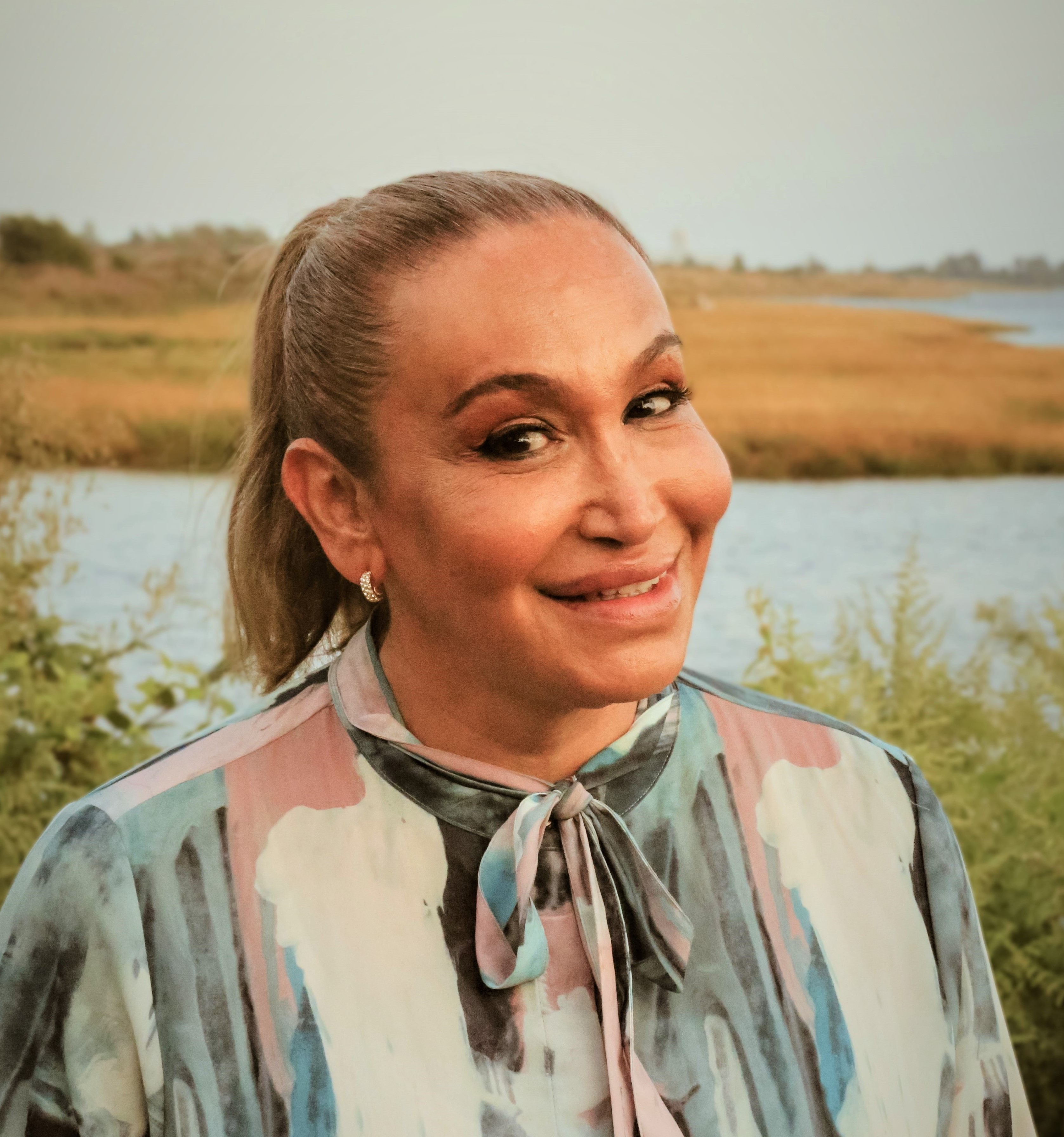 Cecilia, a gorgeous latina transsexual woman, stands outside in front of a body of water, surrounded by plants. She wears a multicolored pink, black, blue and white blouse with a bow tied around her neck. She looks directly in the camera and smiles.