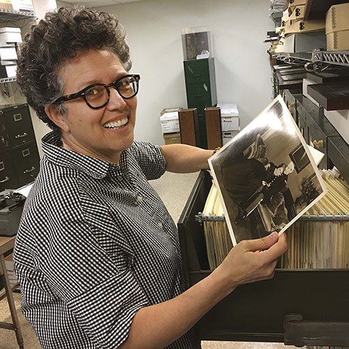 Chana Pollack is wearing a short sleeved black and white checked button down, black thick-rimmed glasses, and is standing in front of a file cabinet, holding a photograph, and looking at the camera and smiling.