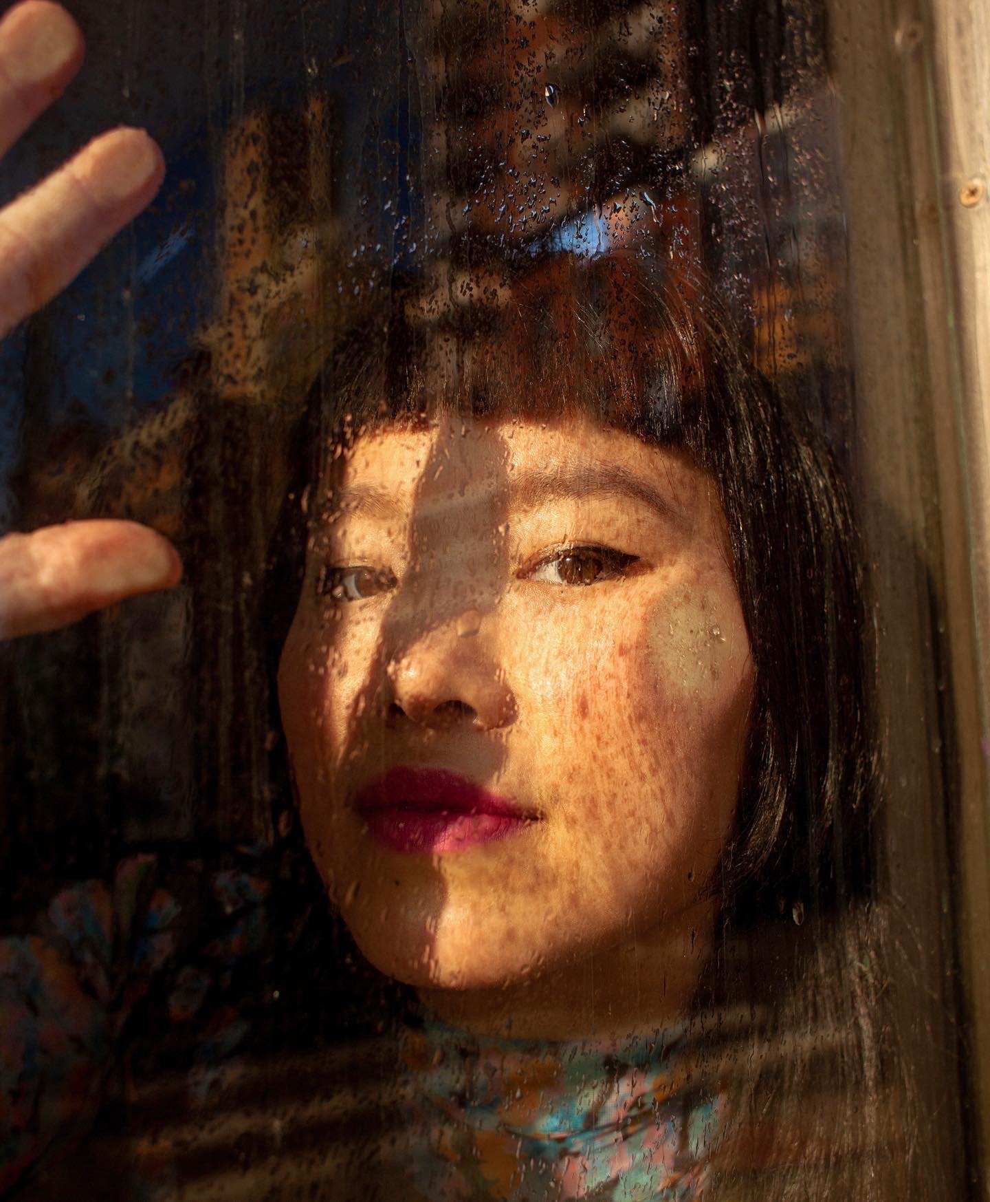 Charmaine Lee is photographed through a window, her cheek and fingers touching the glass, looking at the camera