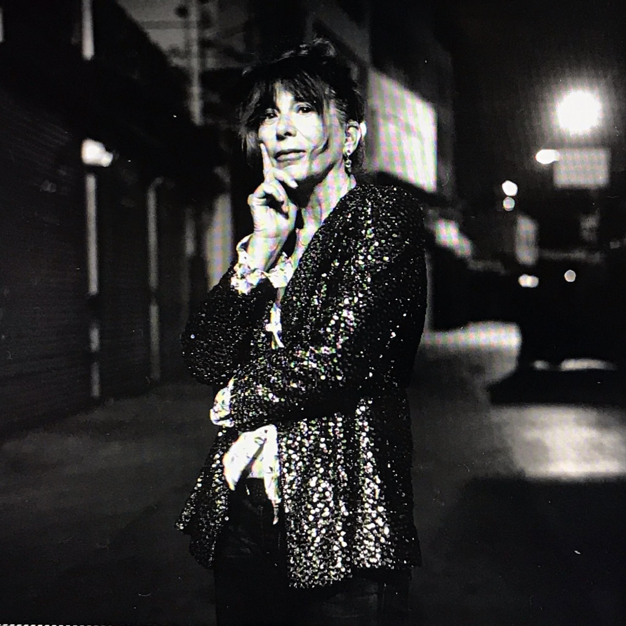 In this black and white portrait, Chris Kraus wears a sparkly jacket and looks at the camera with her face in her hand