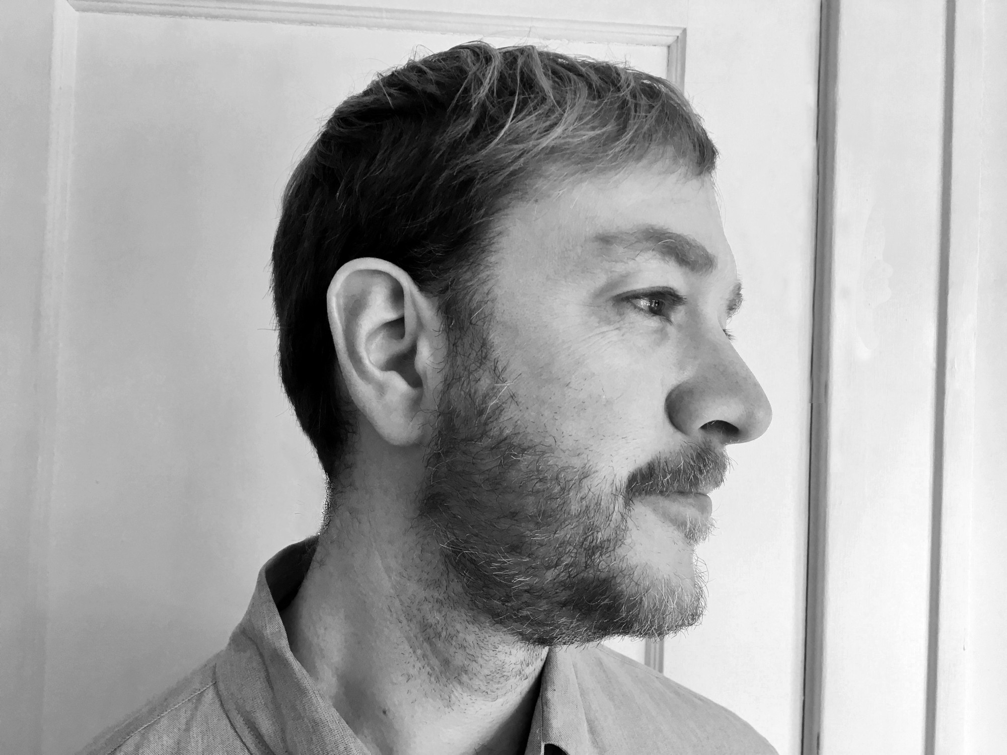 Chris Martin, captured here in black and white, offers you his right ear. He is a white man with short hair, a multicolored beard, and a collared shirt.