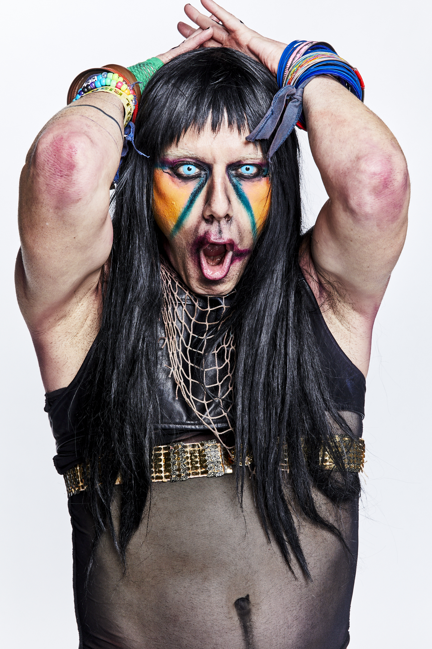 CHRISTEENE is shown from the waist up with her arms up, elbows out, and hands on the back of her head. She is open mouthed, very wide eyed, and is emitting some kind of sound from her mouth. Her face is painted with vibrant colors which are geometrically shaped below her eyes in a triangle pattern. She has long black hair and piercing blue eyes. She appears dirty as in not clean.
