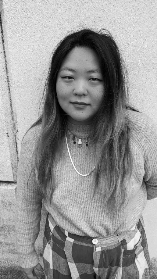 Christine Kwon is featured in a black and white photograph in portrait-format, cropped from the waist up. She is wearing checkered pants, a sweater, a long chain necklace and a shorter chain necklace with droplet pendants. Her hair is long over her shoulders and she is looking directly into the camera. Behind is a plain cement wall. The photograph is tightly cropped but she appears to be outdoors.