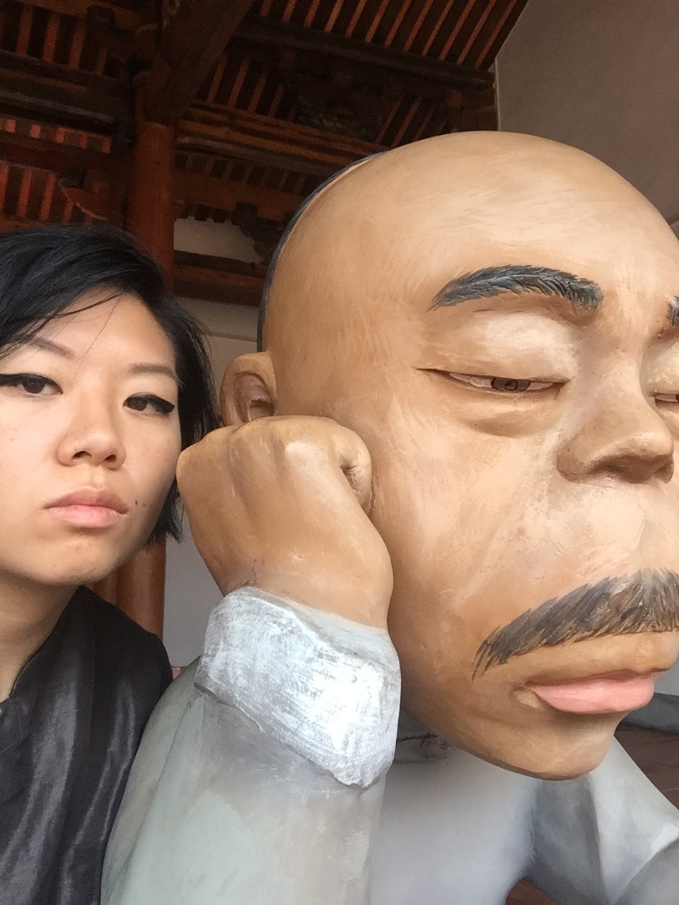 Cindy Gao is to the left of the image, facing the camera. Cindy is standing next to a sculpture of a bald man with a moustache, resting his cheek on his fist.