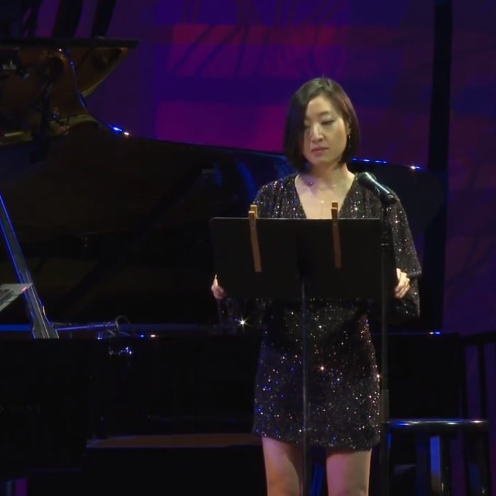 Cindy Tran stands on a stage at music stand in front of a grand piano, wearing a black sparkly dress