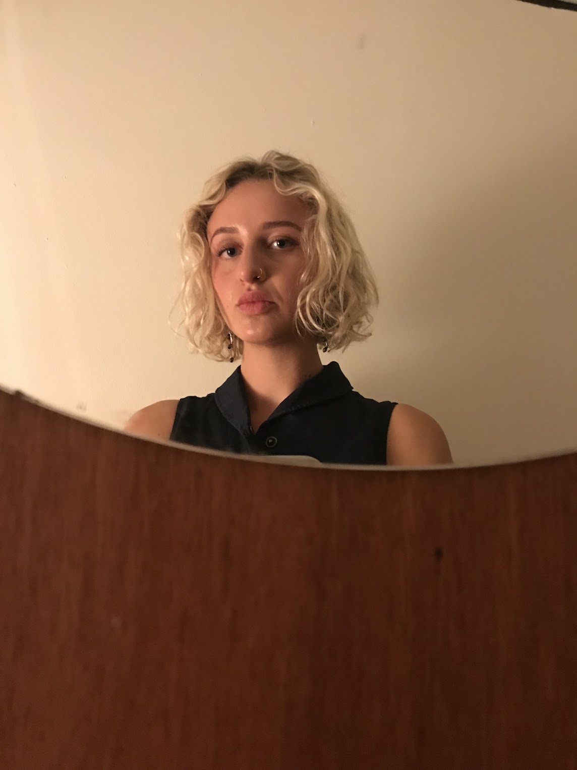 Clementine in a dark vest stands in front of a mirror mounted on a wooden wall.
