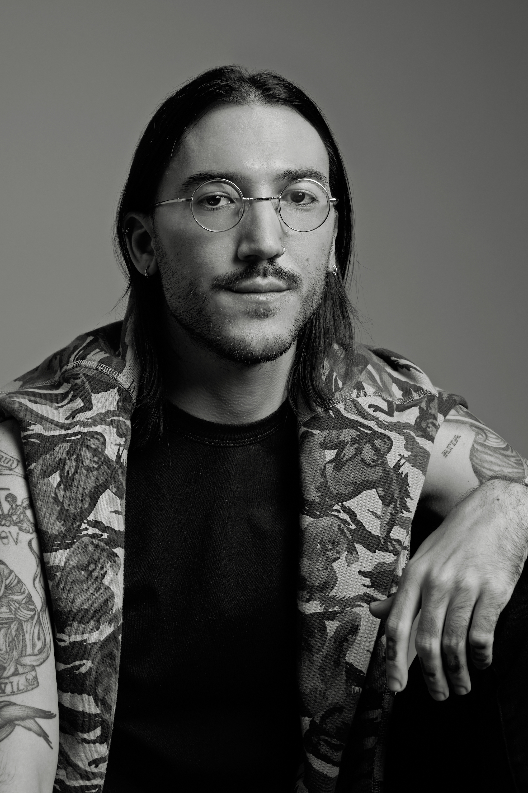 Black and white photograph of Constantine, a white person with shoulder-length black hair, round glasses, and silver earrings, wearing a black shirt with a camouflage print sweater draped over their shoulders.