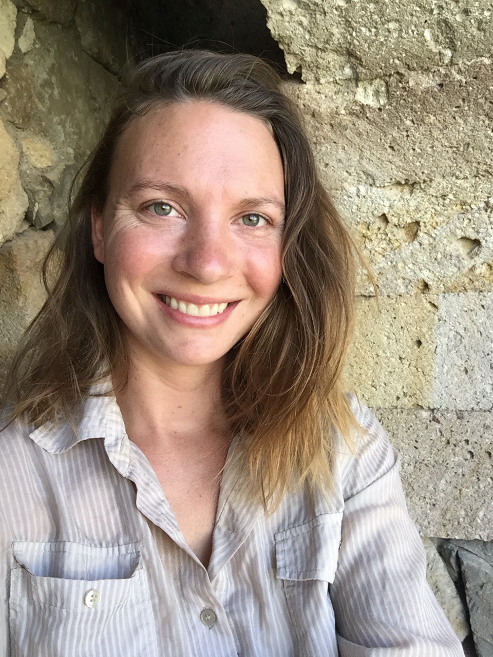 Corrine Beardsley stands in front of a stone wall wearing a linen button up and smiling at the camera