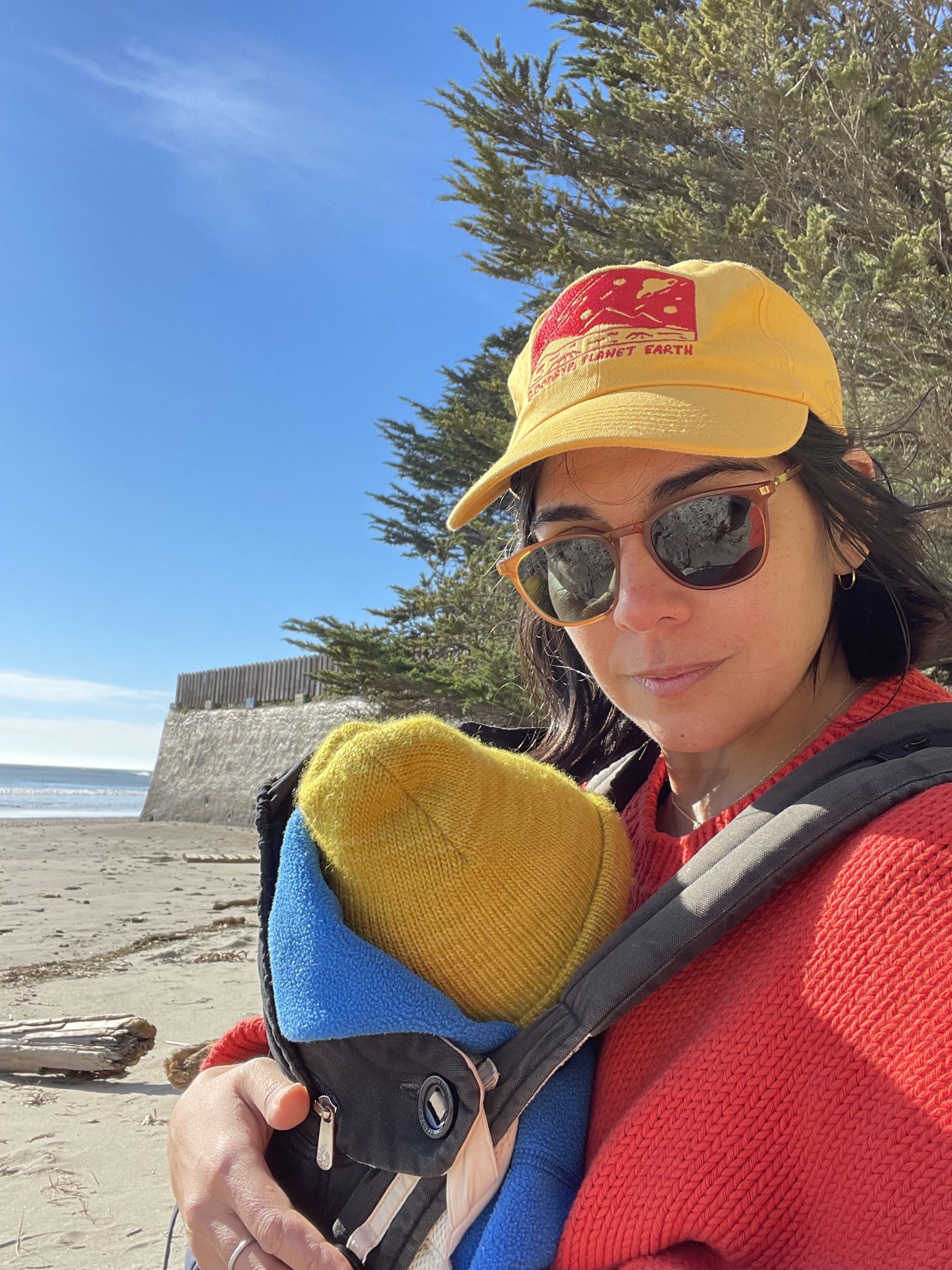 Corrine Fitzpatrick is sitting at the beach under the blue sky, wearing a red sweater, sunglasses, and yellow baseball cap. She is holding a baby wearing a mustard yellow beanie, who faces away from the camera and towards Corrine.