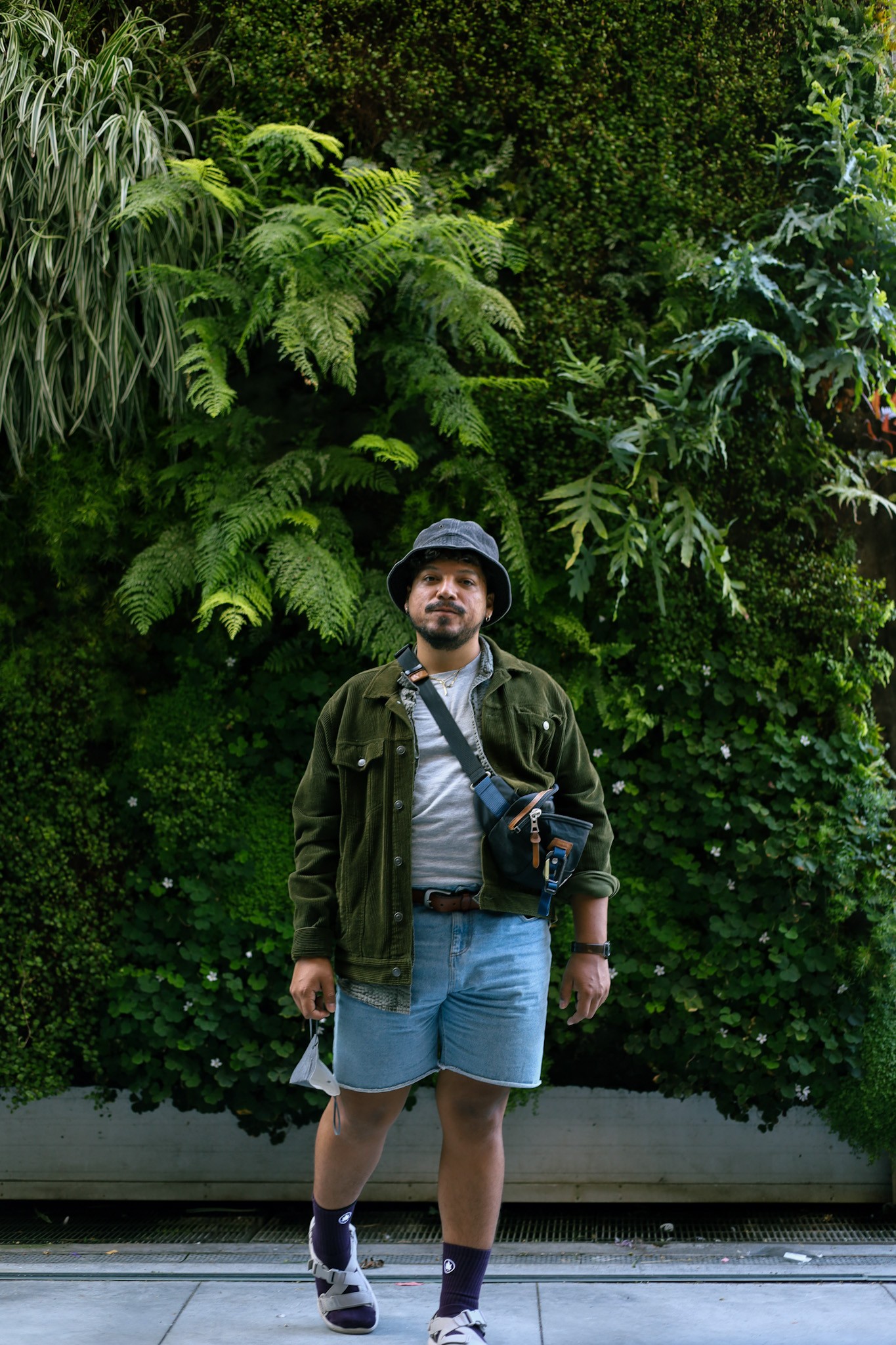 Cristóbal Guerra stands in front of a wall of verdant foliage, looking into the camera, wearing a blue bucket hat