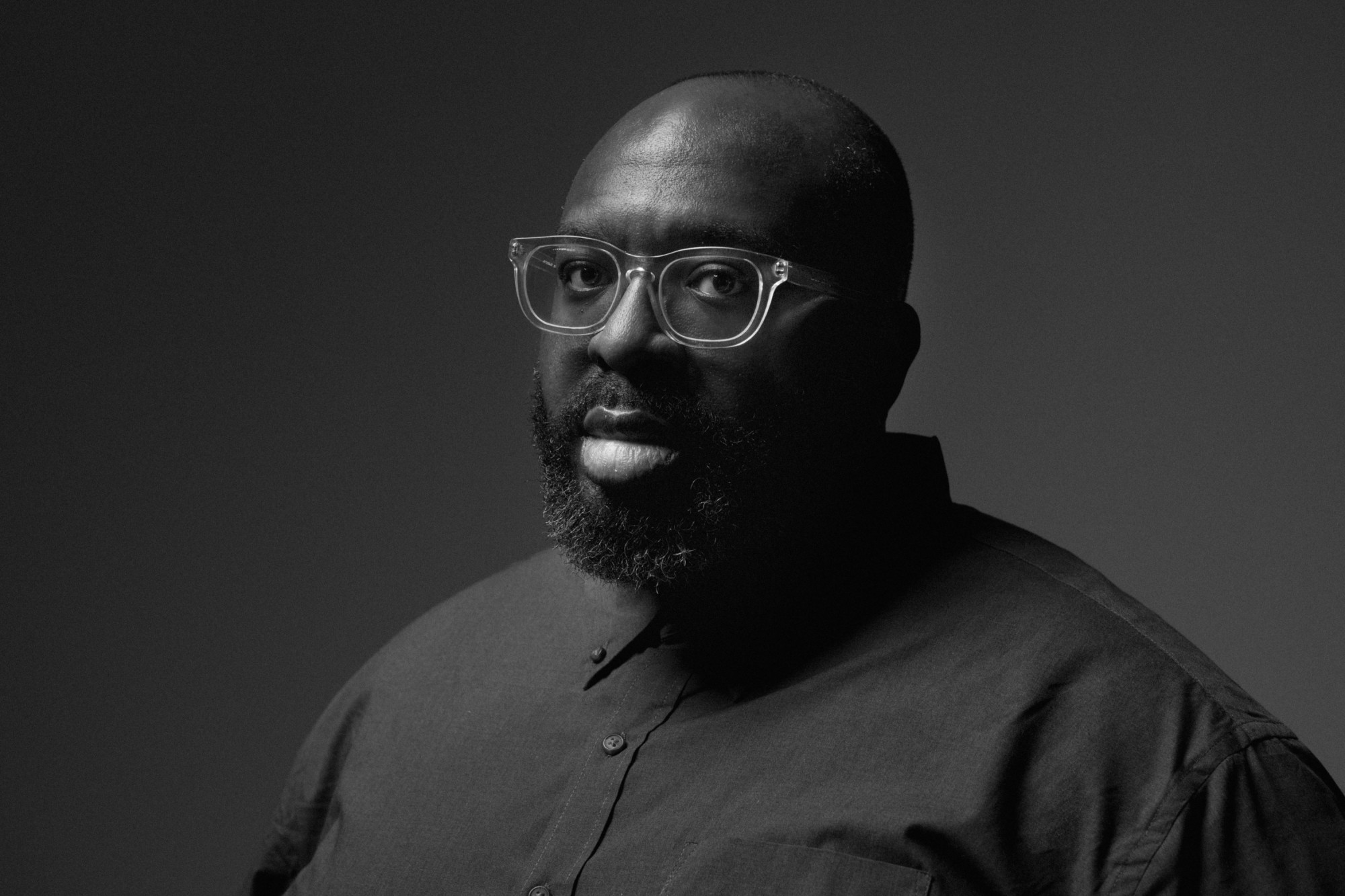 Darius Jones is featured center in a black and white landscape-formatted portrait. He is seated in front of a black backdrop wearing a button down and glasses, facing the camera. The picture is cropped from the shoulders up.