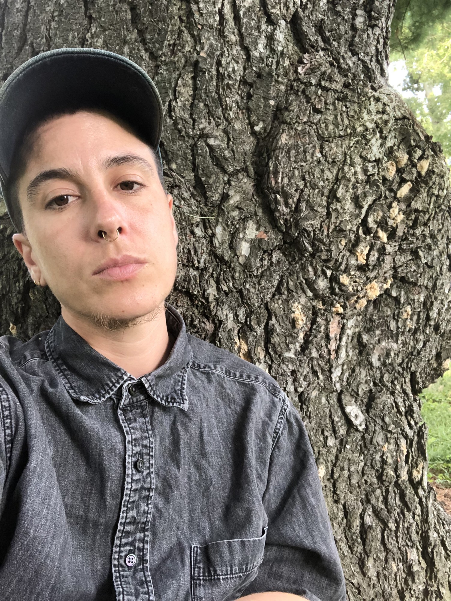 devynn emory is shown in a selfie-mode portrait, in the left of the frame, cropped from the torso up, wearing a grey denim button down and leaning against a tree.