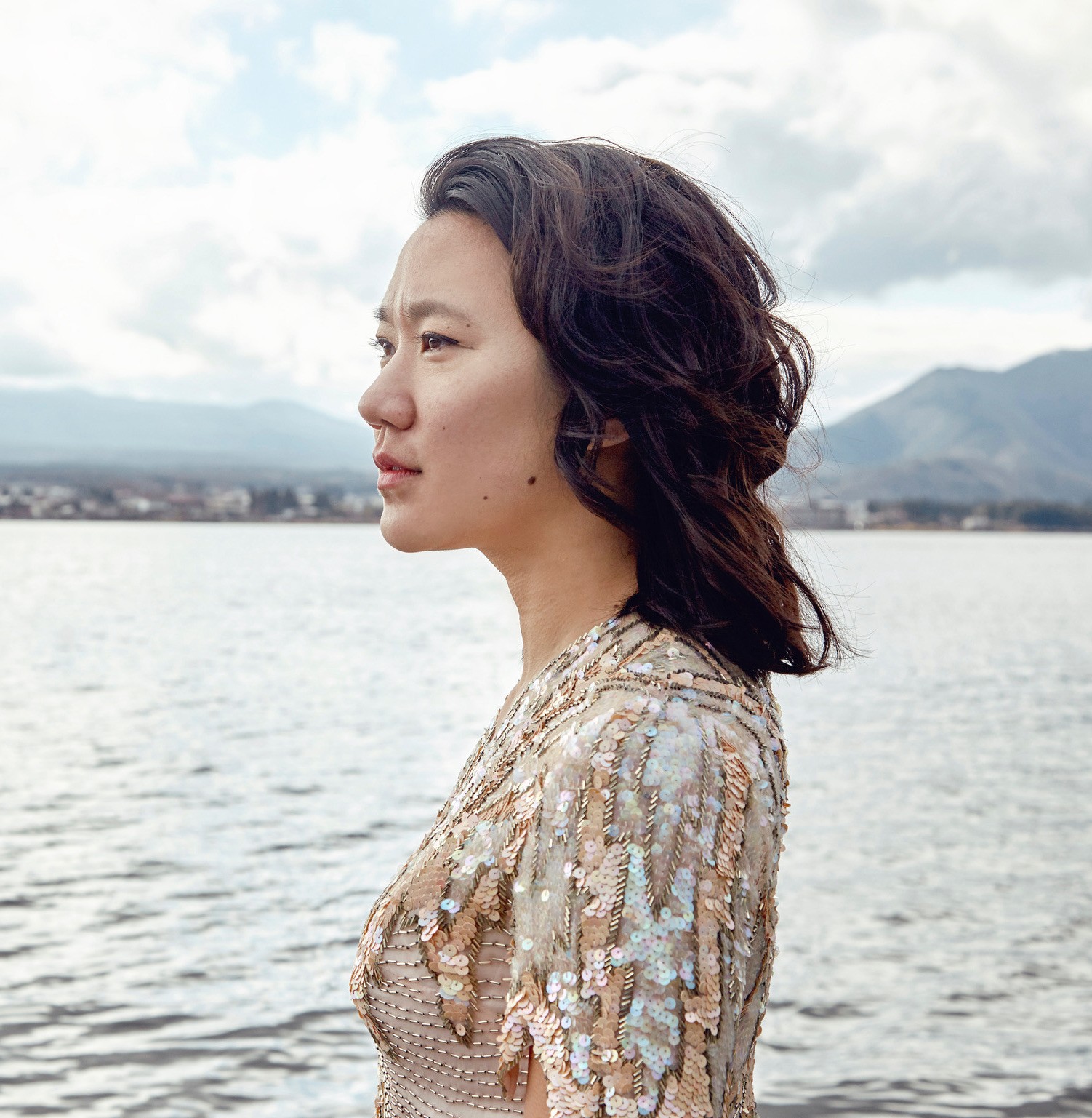 Diana Khoi Nguyen wears a sequined shirt, standing next to a body of water, looking into the distance