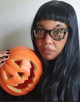 Dianca London Potts looks directly into the camera, holding a jack-o-lantern. Around her left eye are smaller eyes photoshopped into the image. She wears black enamel glasses and Bettie Page bangs.