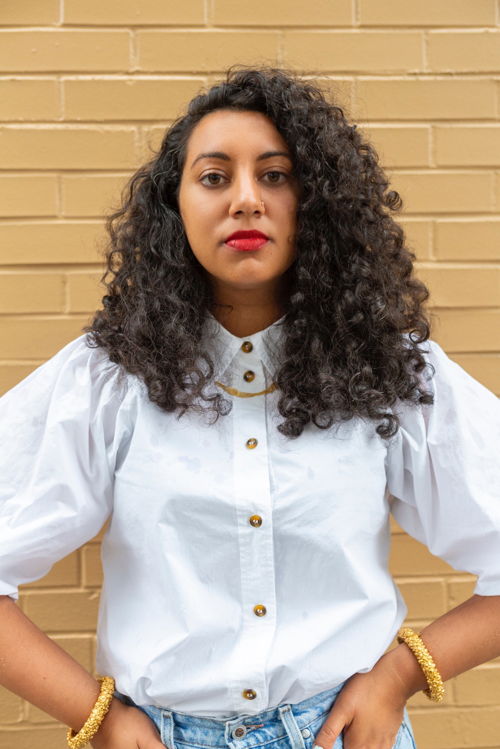 Diya stands, arms at hip, against a yellow-ish brick wall. She wears a white button down shirt with big sleeves, gold bangles and hoops, and a red lip.