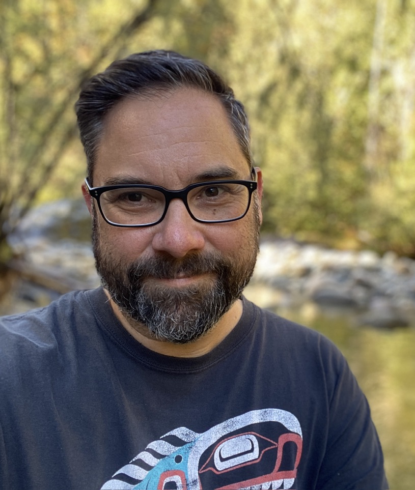 A man with dark hair and black-rimmed glasses, sits in front of a river and sun-filled forest, smiling. He is wearing a shirt with See Monster image by Wuikinuxv and Klahoose Nation artist Bracken Hanuse-Corlett.