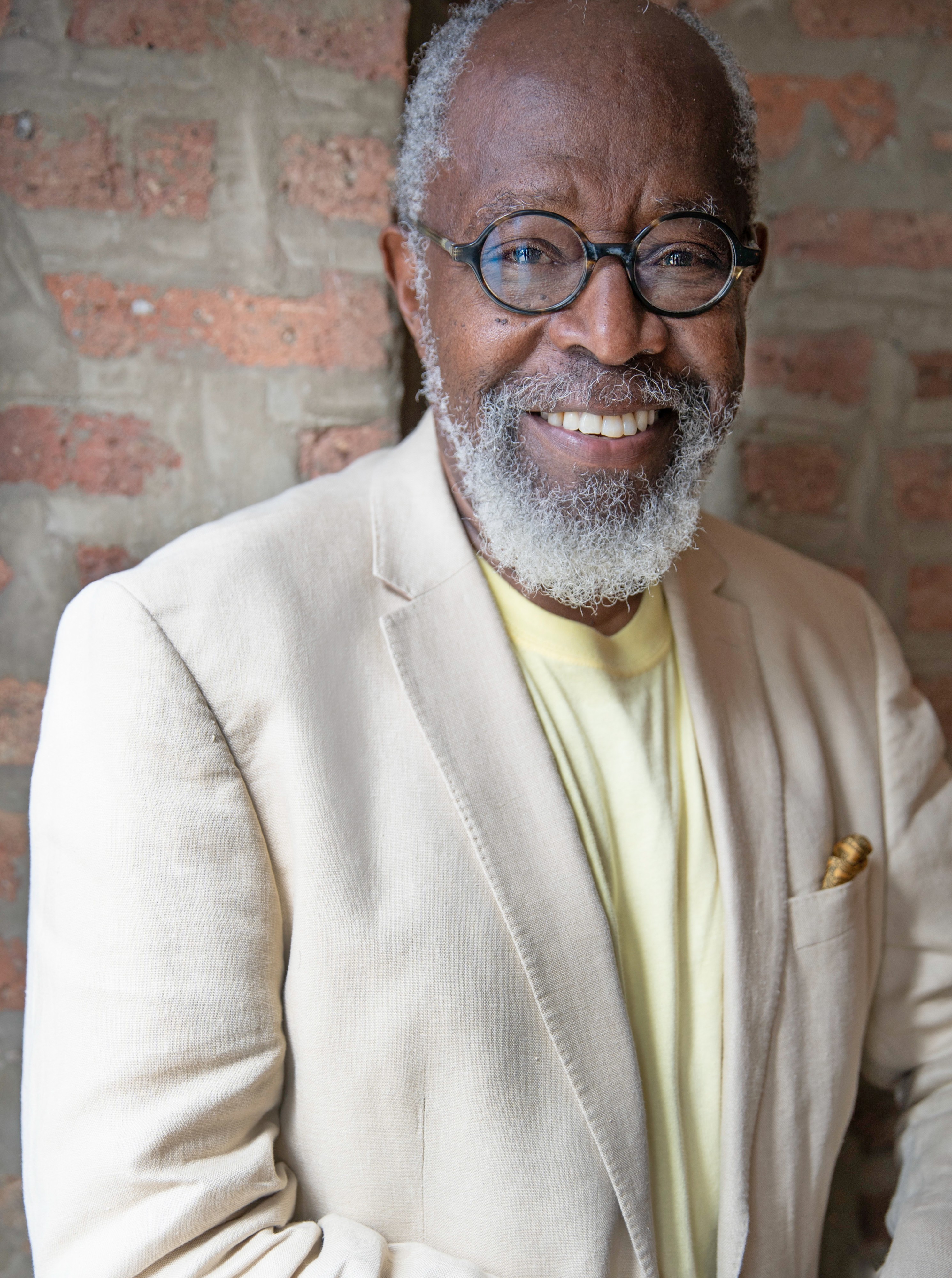 Ed Roberson wears a pale jacket and light yellow shirt, sitting in front of a brick wall, smiling and looking at the camera
