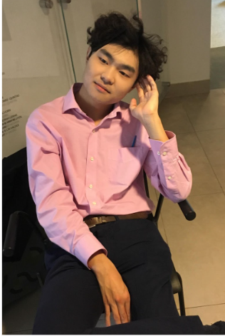 Eddy, a Chinese man with dark hair, sits in a black chair atop ivory tiles, surrounded by a white pillar and glass paneling. He is wearing a pink button up shirt, dark slacks, and a brown belt. His hand rests against his left ear. He looks to the left of the frame at an angle, with a smile.