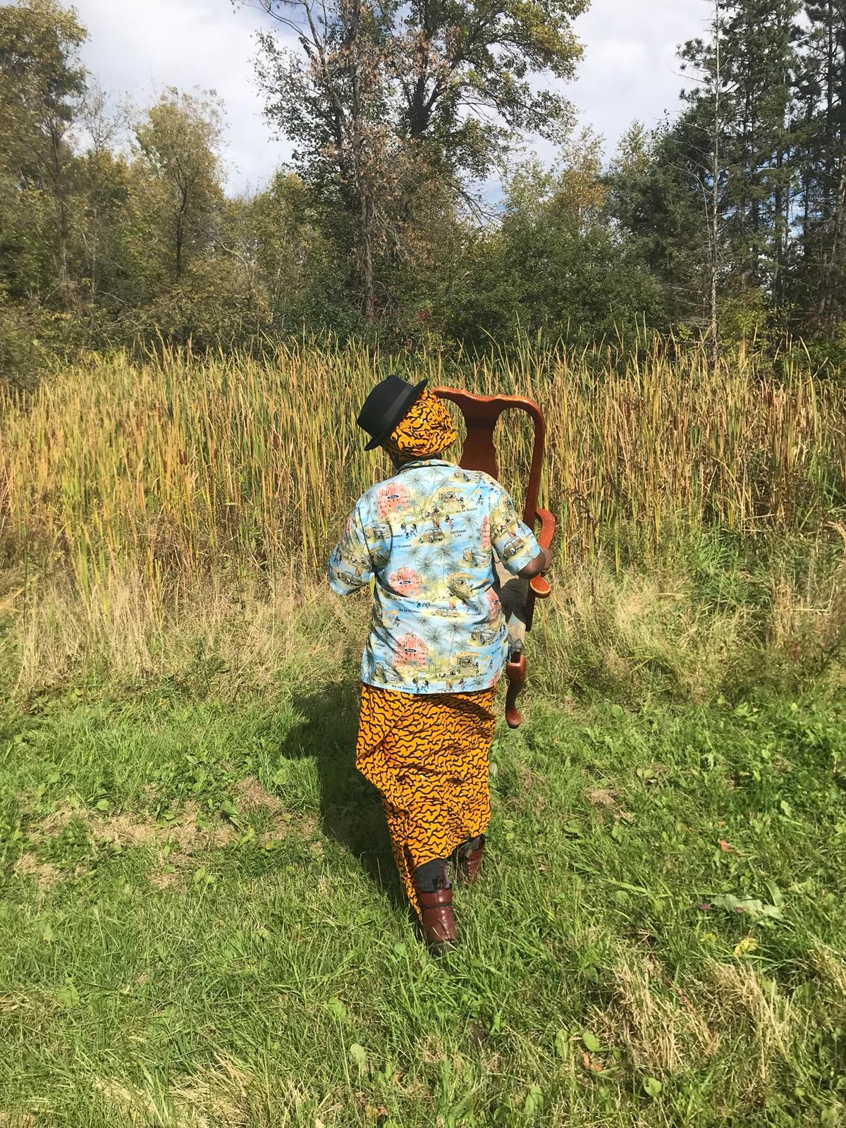 Ego Ahaiwe Sowinski is facing away from the camera, towards tall grass and trees, carrying a wooden chair. She is dressed in brightly patterned fabrics, and wearing a fedora on top of a headscarf, tilted at an angle.