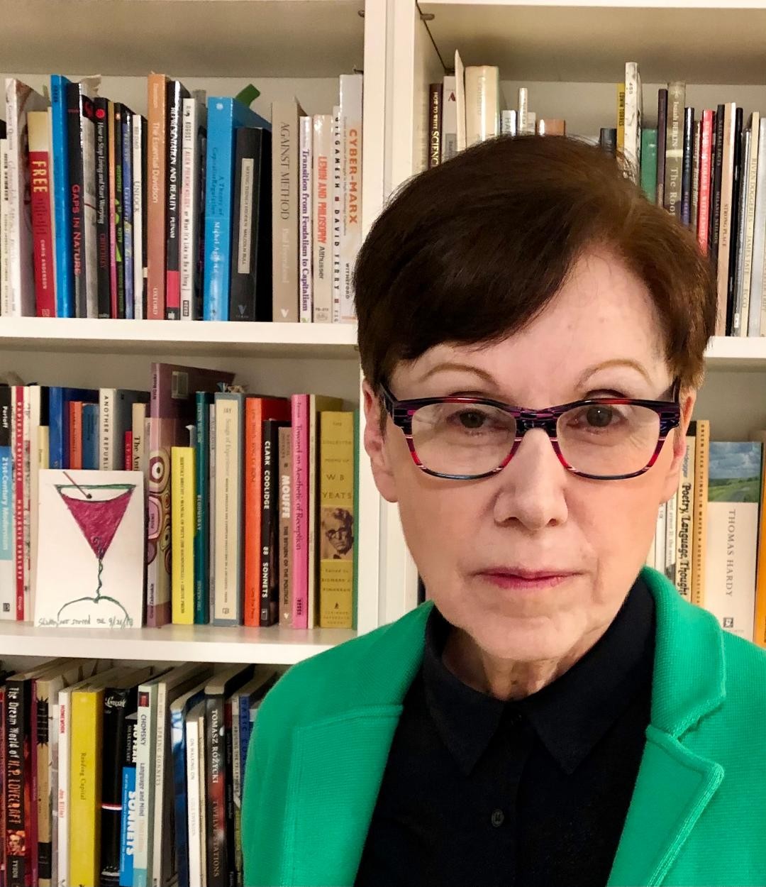 Elaine Equi stands in front of a bookshelf, wearing a black button-up and green blazer.