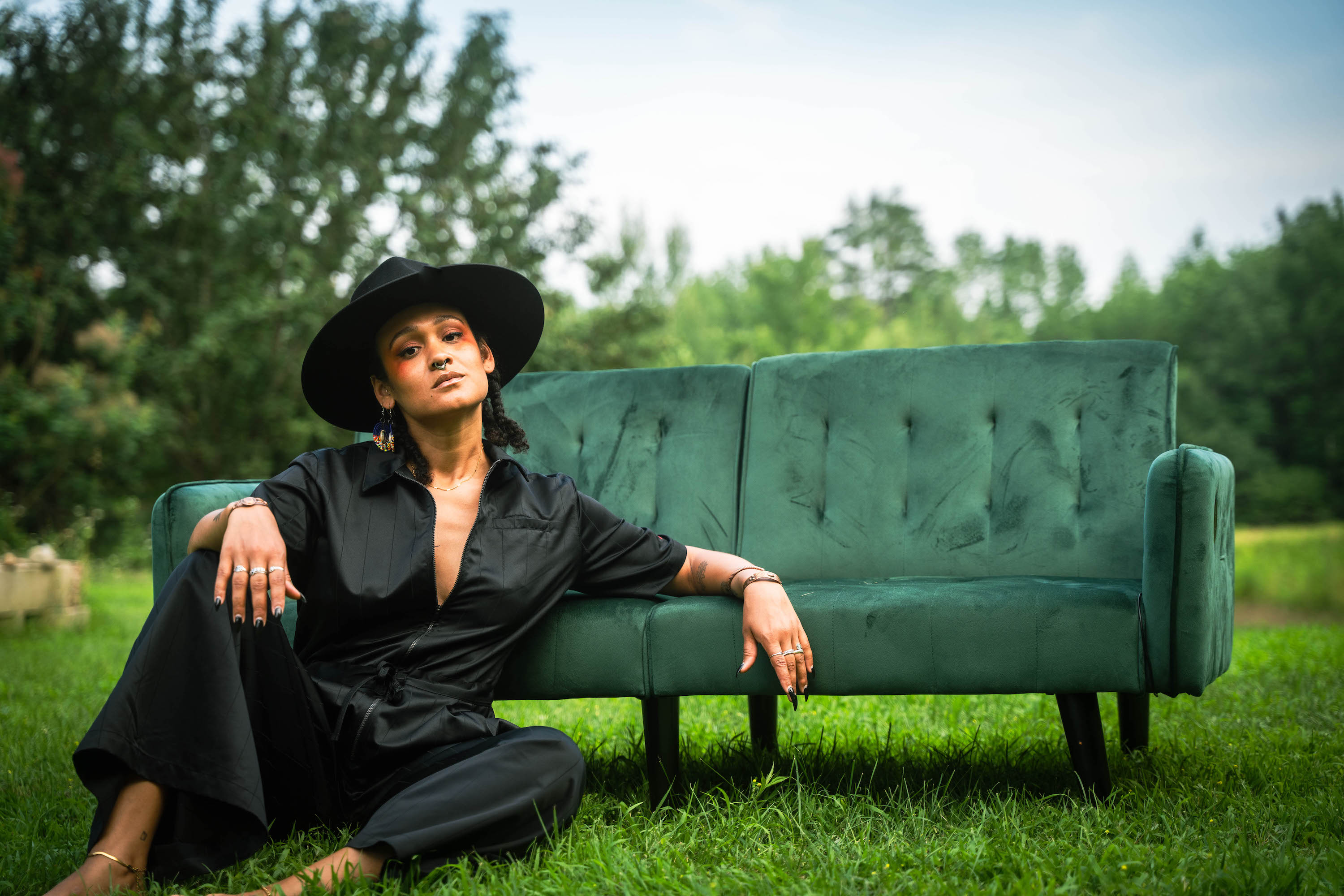 Eleanor is wearing a black jumpsuit and wide-brim black hat. They are sitting on a large lawn and leaning against a green velvet couch. Their left arm is resting on their knee.