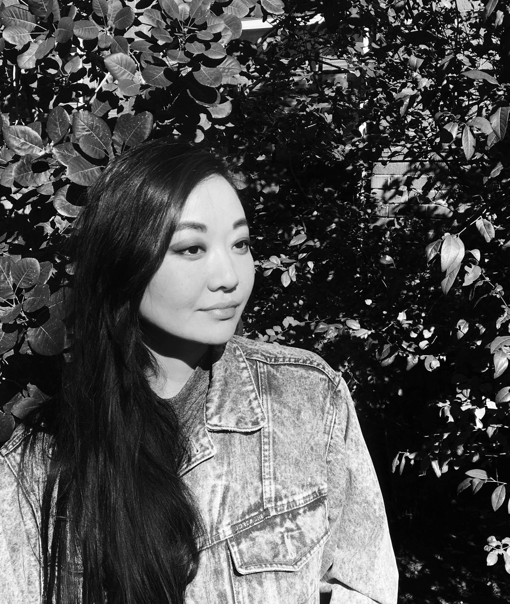 Erinrose Mager is shown in a portrait-oriented black abd white photograph, cropped from just above the waist up. She is wearing a denim jacket, her hair is loose over her shoulder, and she is looking off to the right of the frame. Behind her is tall shrubbery.